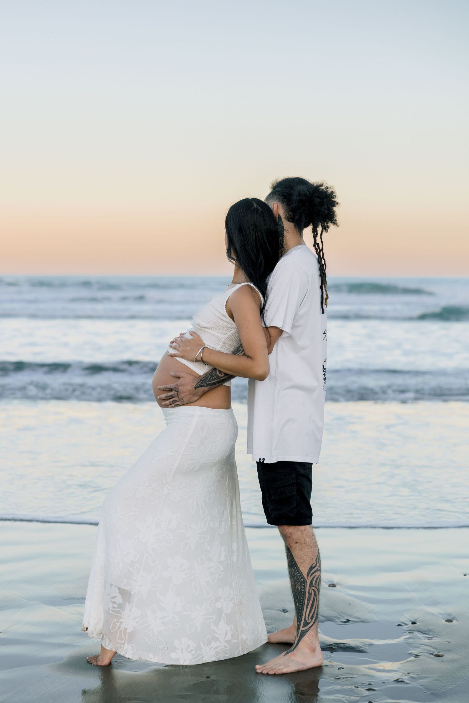 Tattooed couple embracing at sunset beach, man kissing pregnant woman's head near ocean waves.