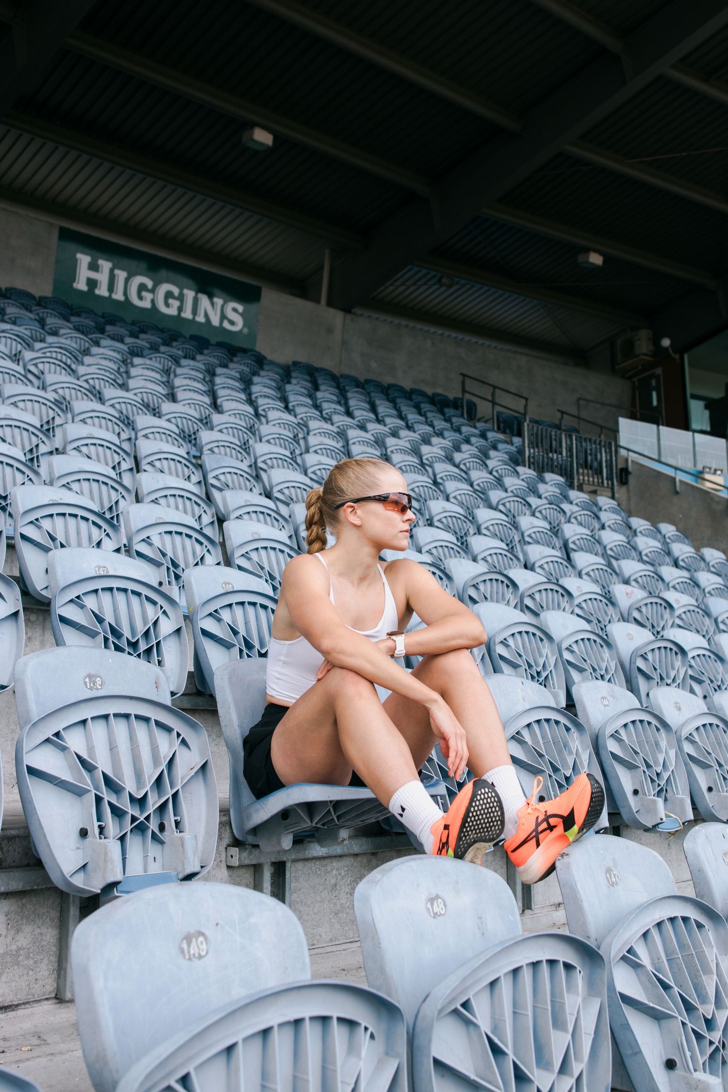 Someone in sunglasses and athletic wear sits alone in empty stadium bleachers at Higgins field.