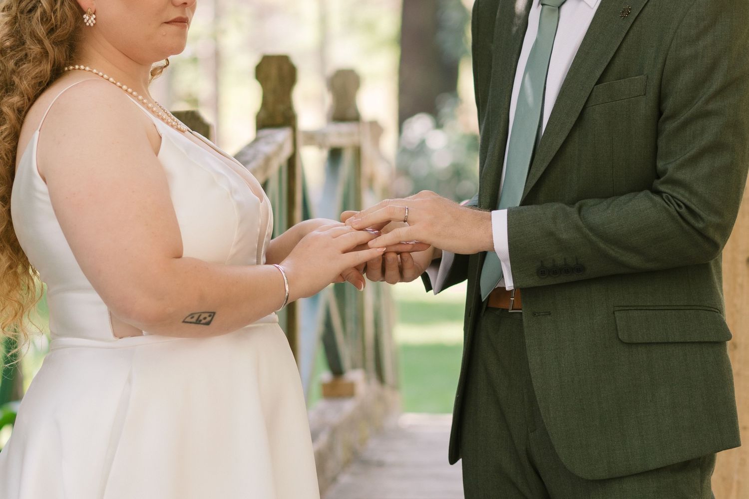 Couple exchanges wedding rings during an outdoor ceremony with person in green suit and white wedding dress.