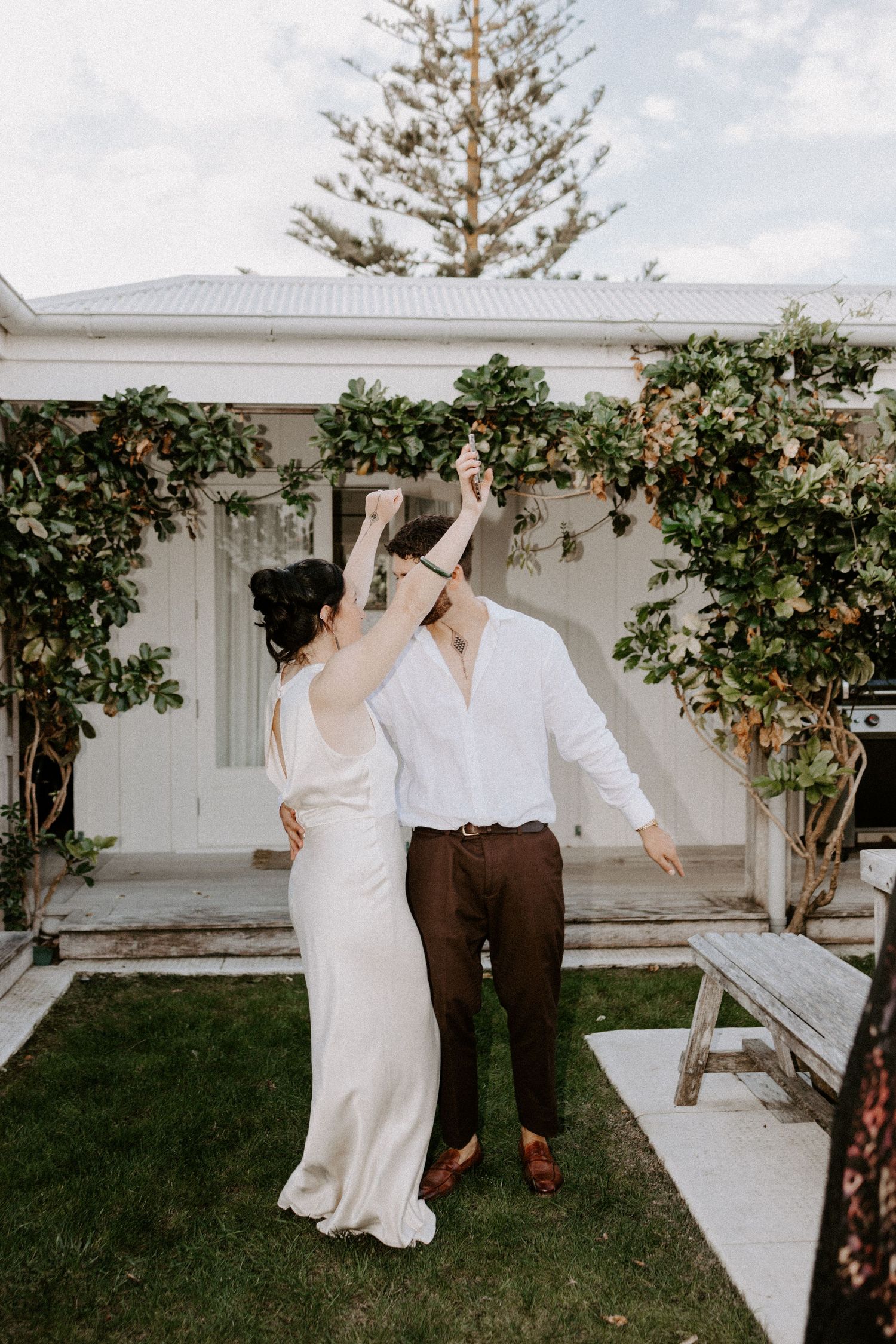 A couple in wedding attire dances in a garden setting with climbing vines and white architecture in the background.