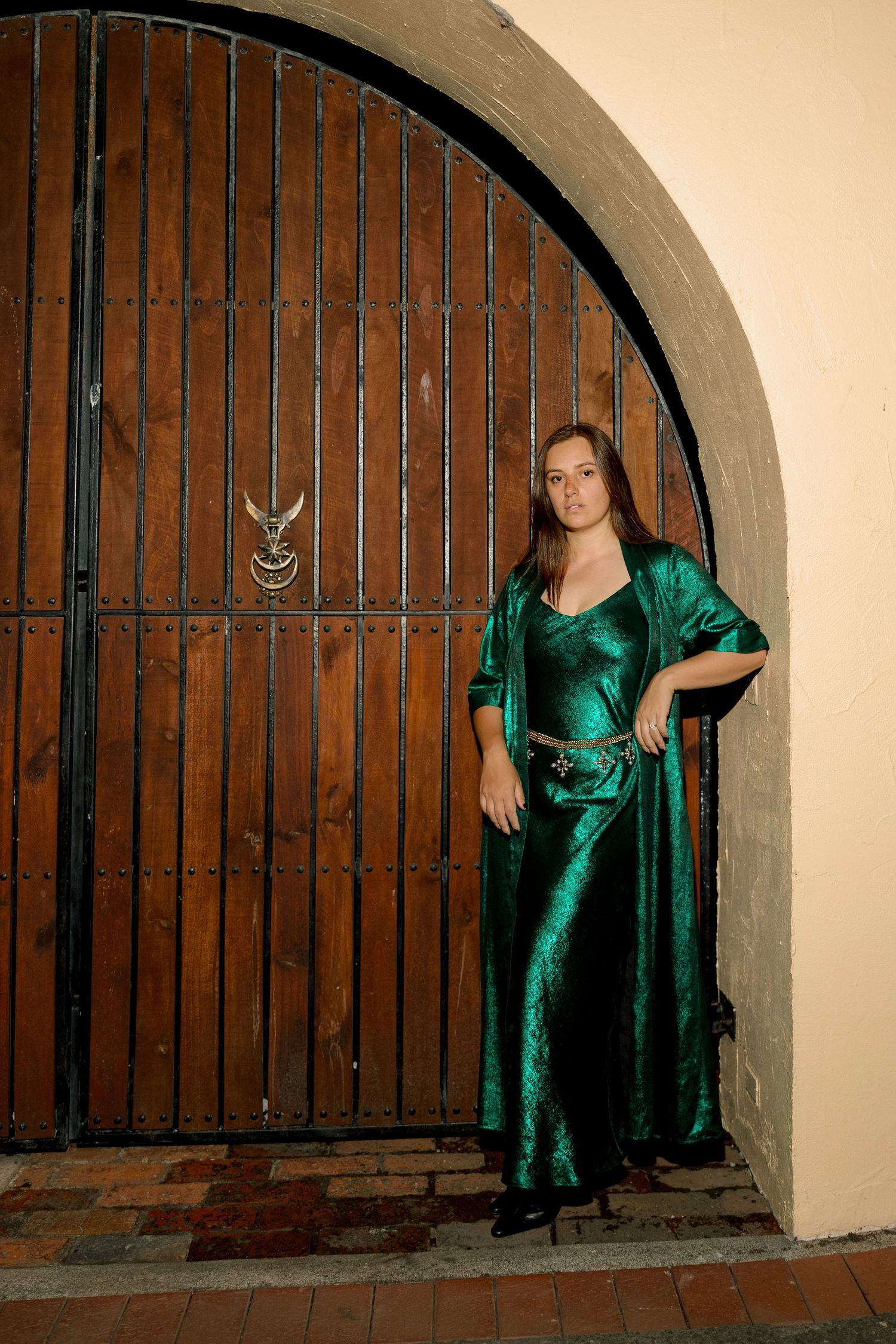 A series of poses in an emerald velvet dress against an arched doorway with vintage wooden gates.