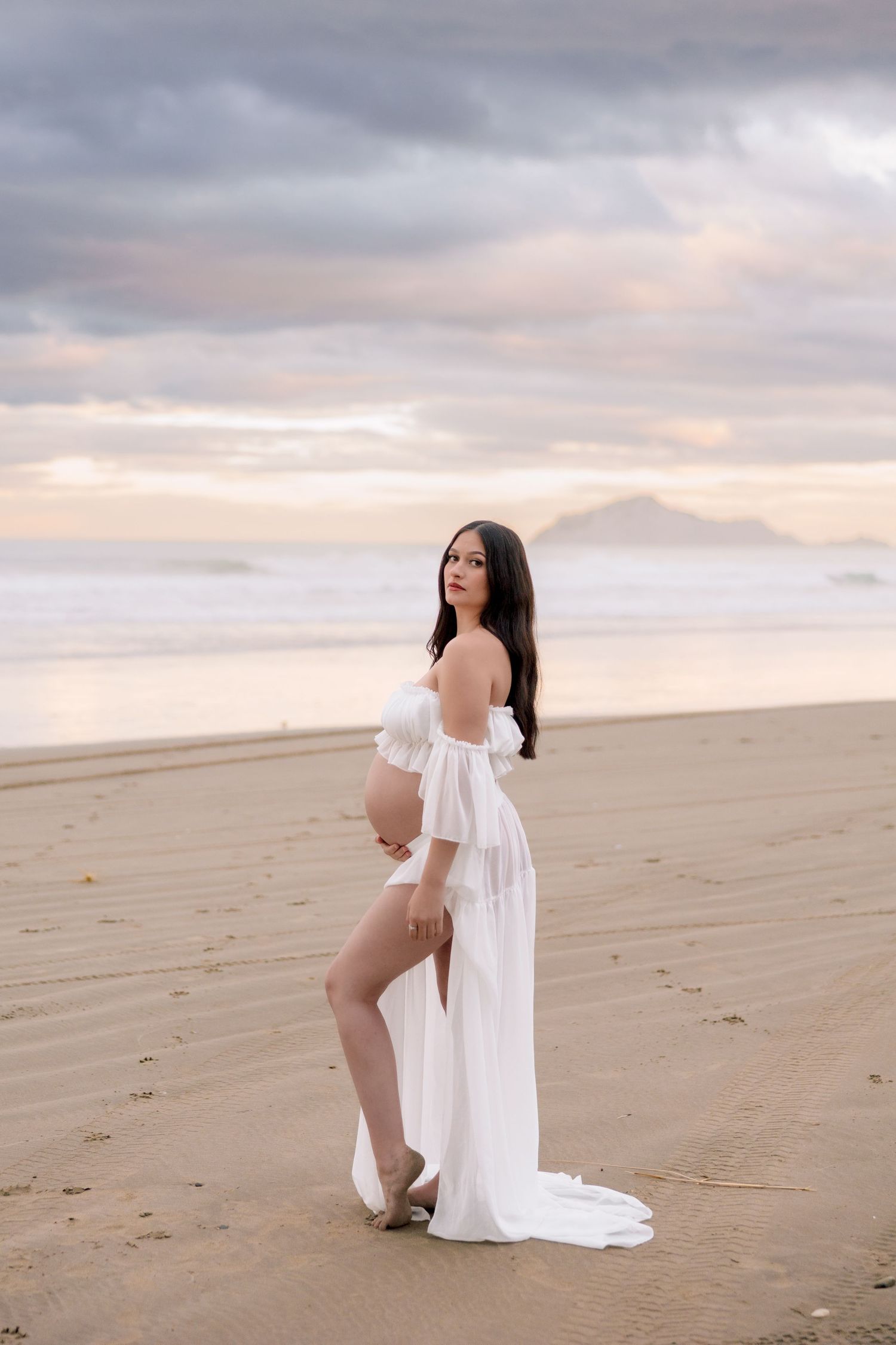 A figure in a flowing white dress poses gracefully on a sandy beach at sunset with dramatic cloudy skies.