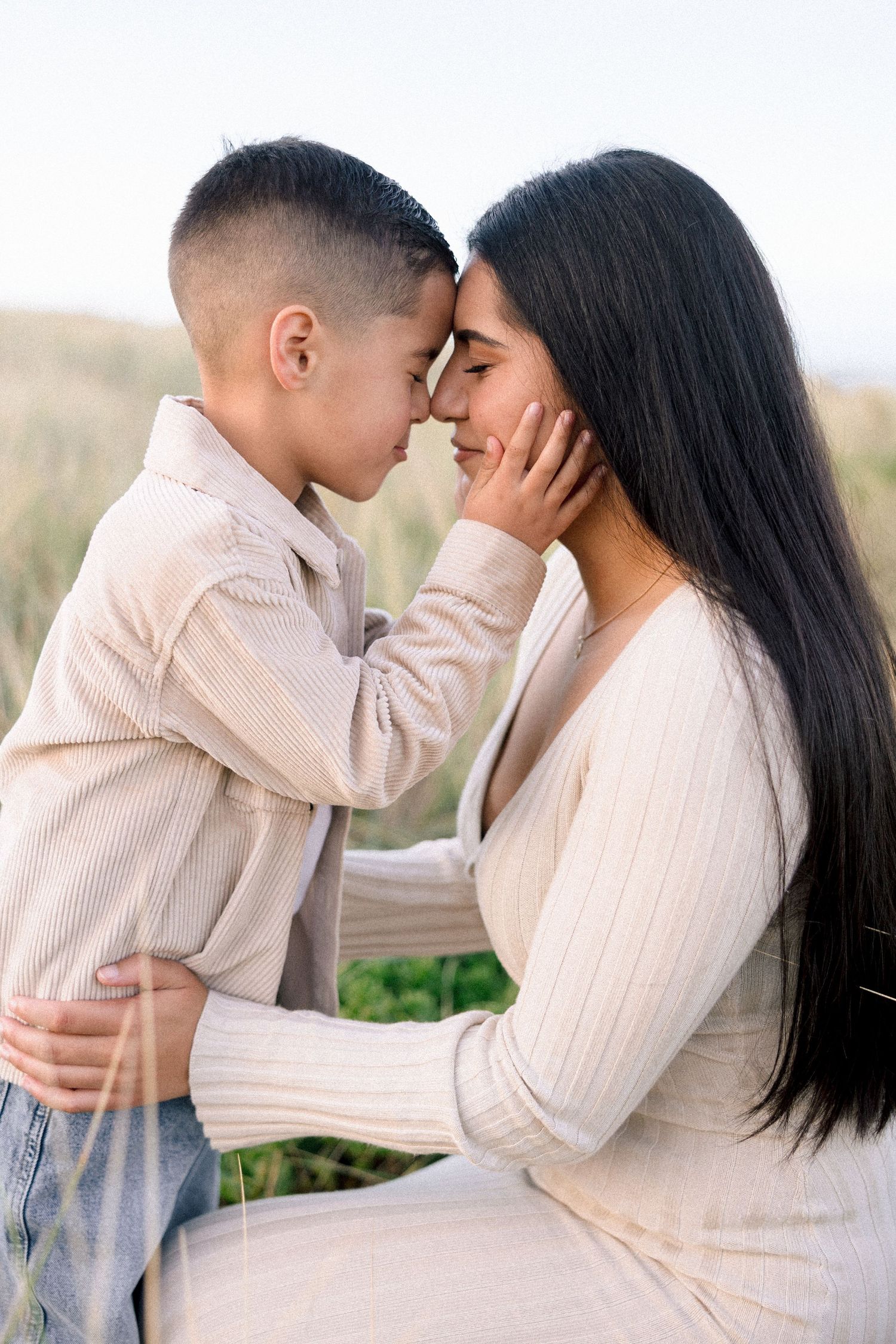 A mother and young son share a tender forehead-to-forehead moment outdoors, both wearing cream outfits.