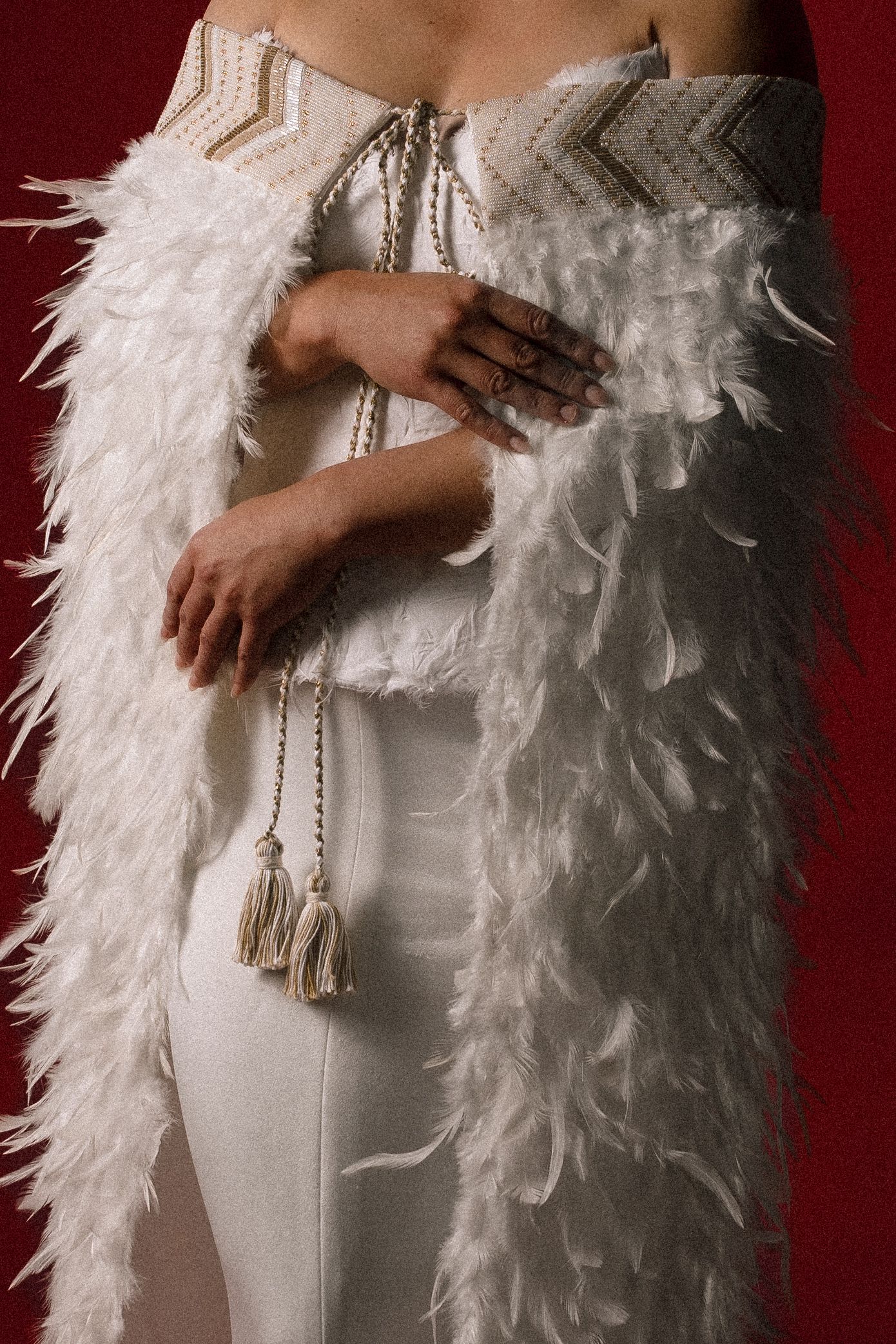 Woman in white feathered wrap and embroidered strapless gown with tassel details on red background.