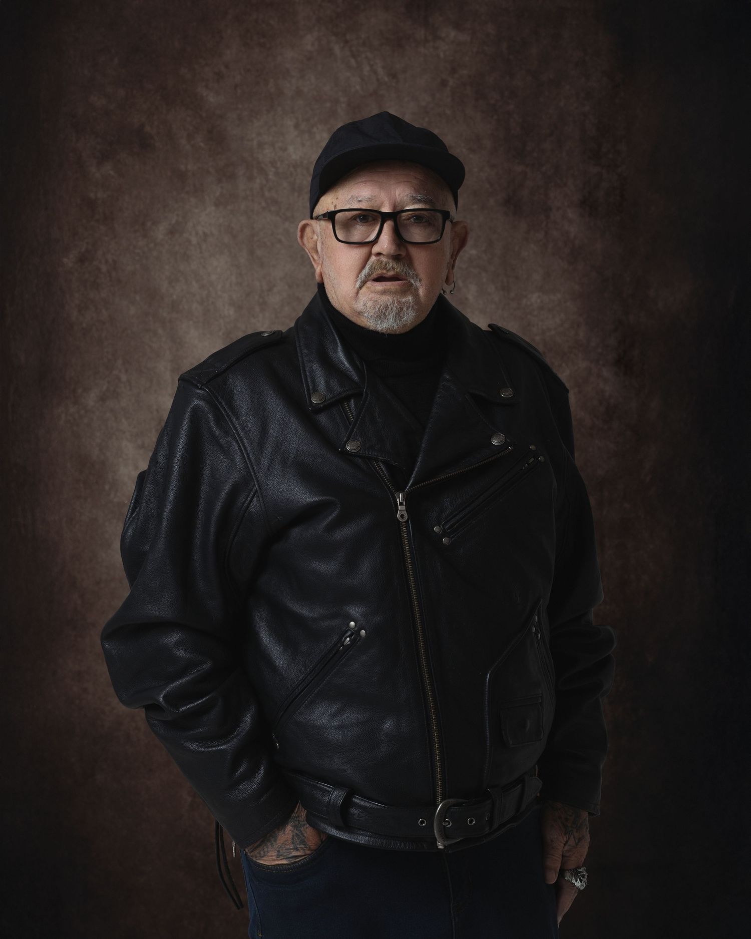 Middle-aged man in black leather biker jacket, cap, and glasses poses confidently against dark studio background.