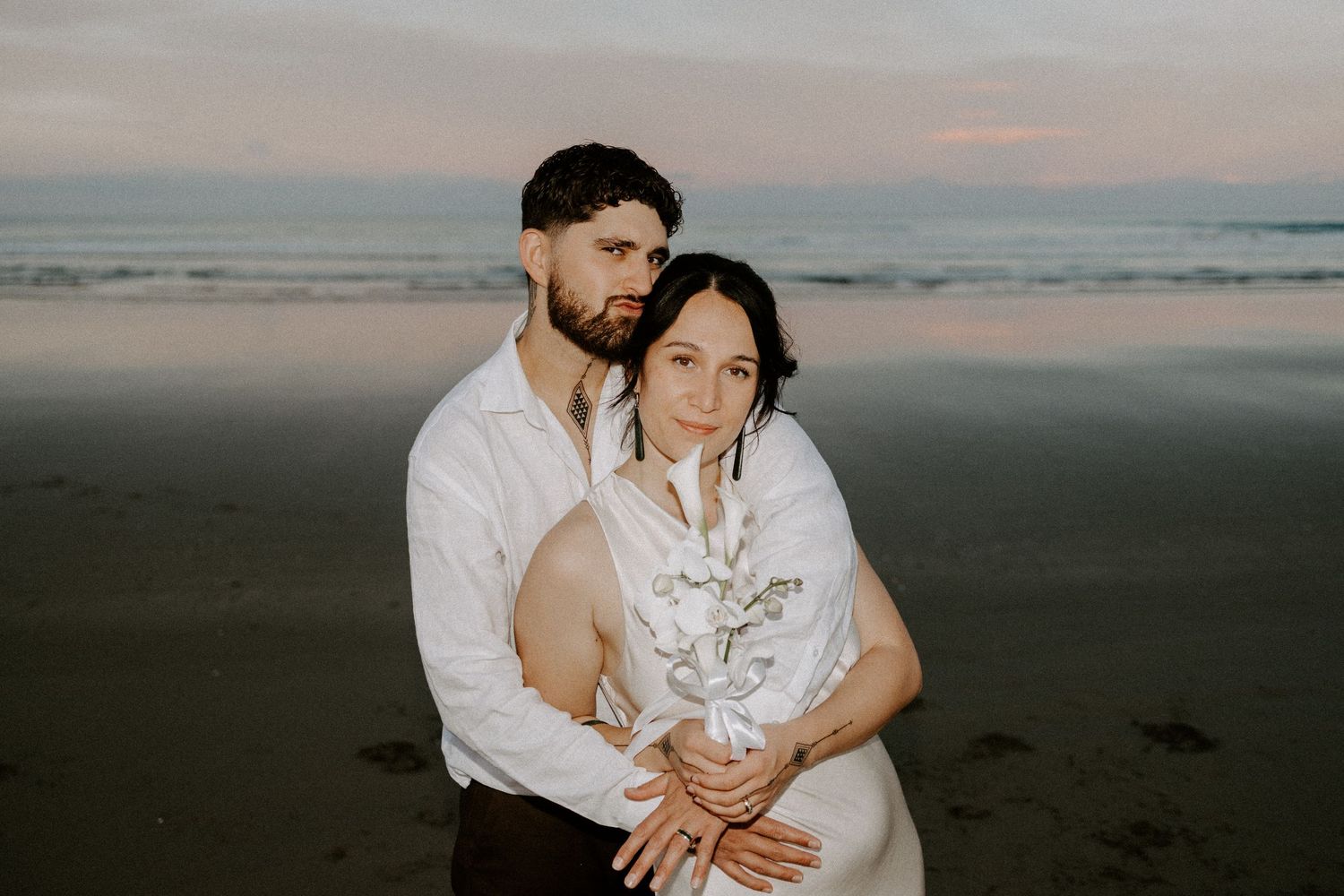 Couple embracing in white outfits during sunset at a beach with waves in the background.