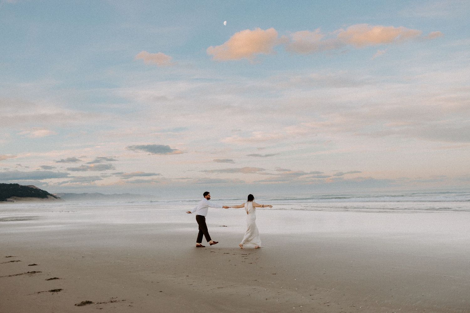 Wide beach landscape with two figures walking hand in hand along the shoreline at dusk.