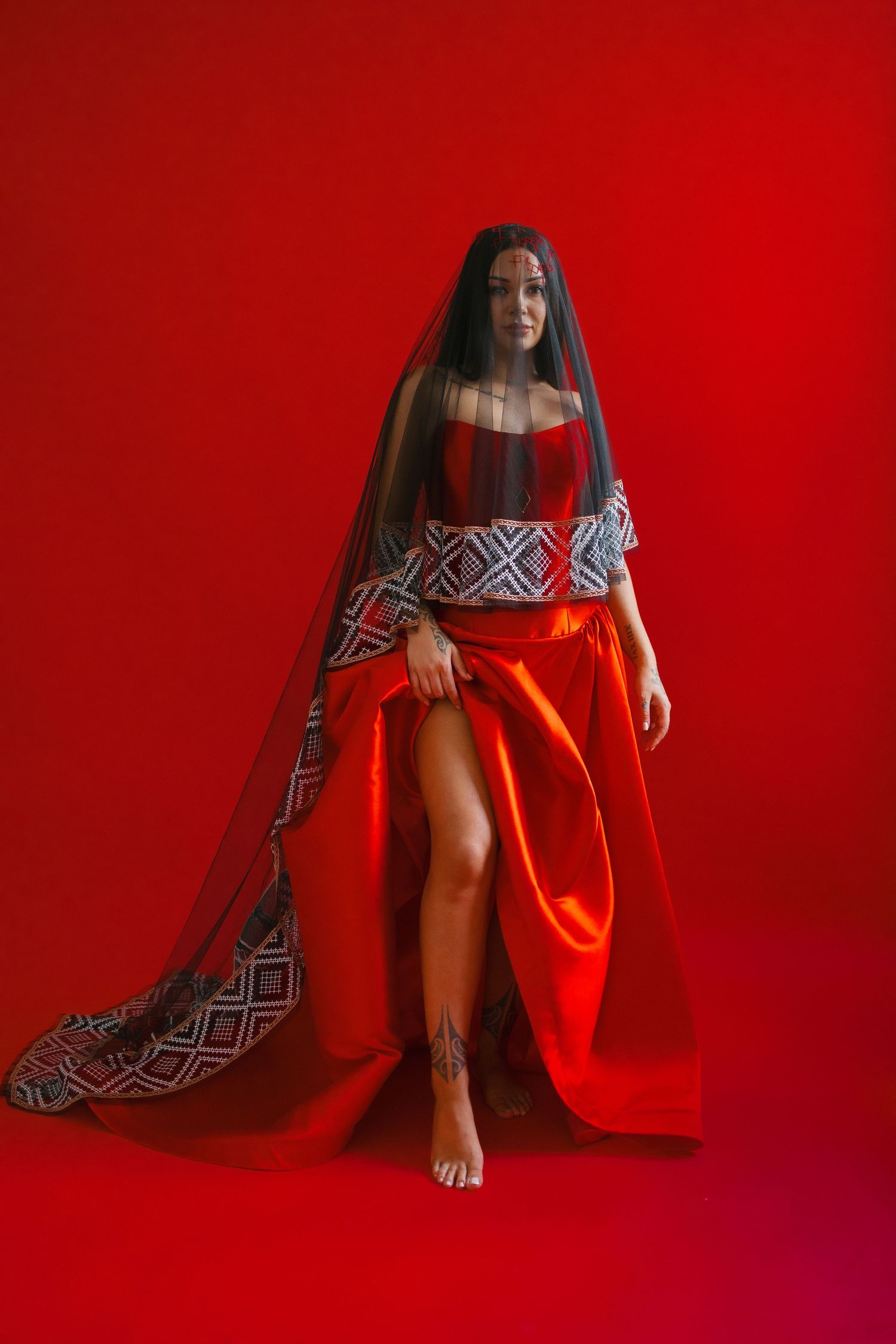 A model in a red dress and long geometric patterned veil poses barefoot against a vibrant red background.