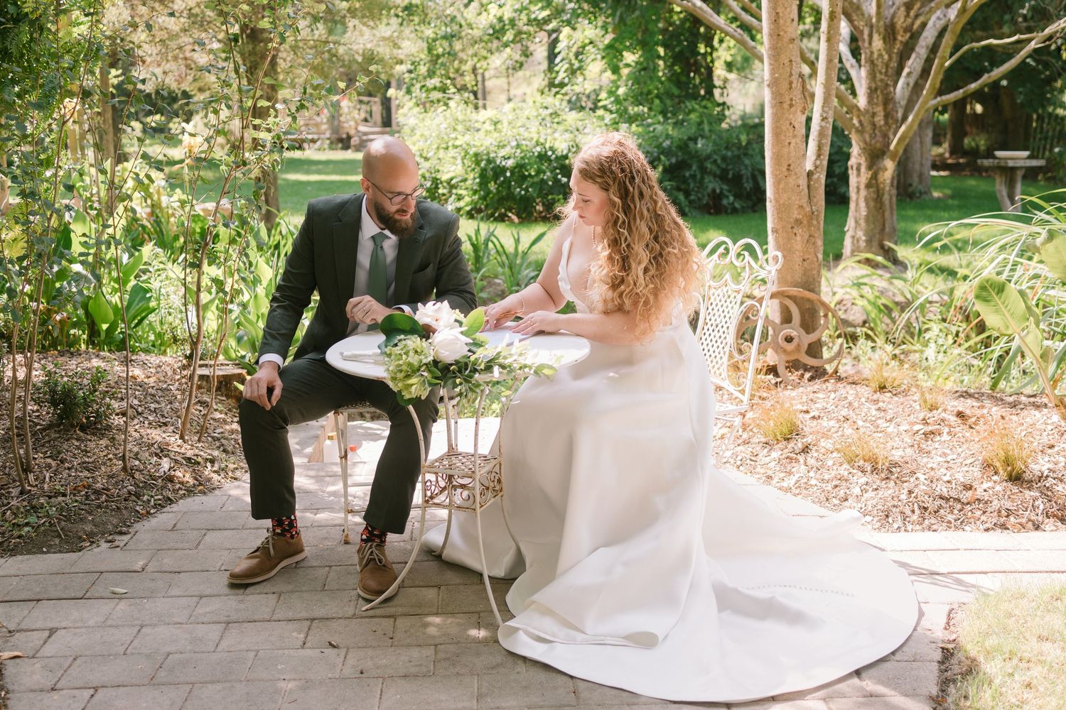 A couple in wedding attire sits at a small outdoor table in a lush garden setting.
