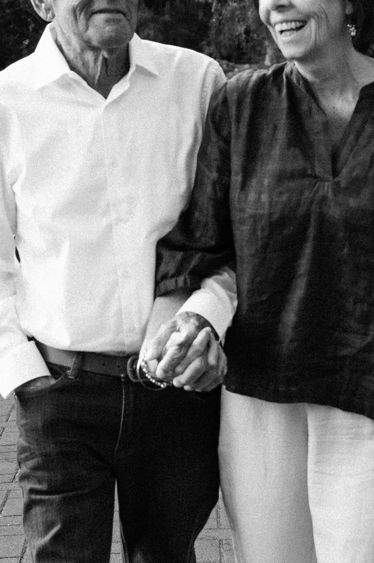 Black and white photo of an elderly couple holding hands, man in white shirt and jeans, woman in dark blouse, smiling warmly.