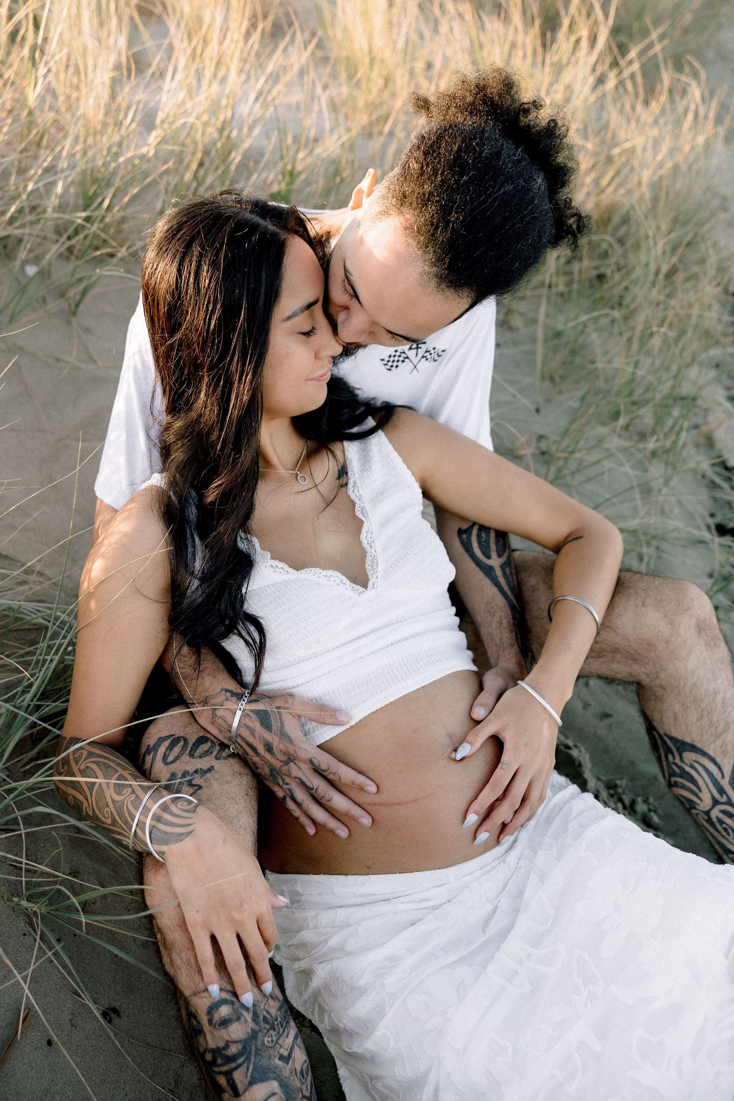 Tattooed couple sharing intimate moment on sandy beach dunes, man kissing pregnant woman in white outfit.