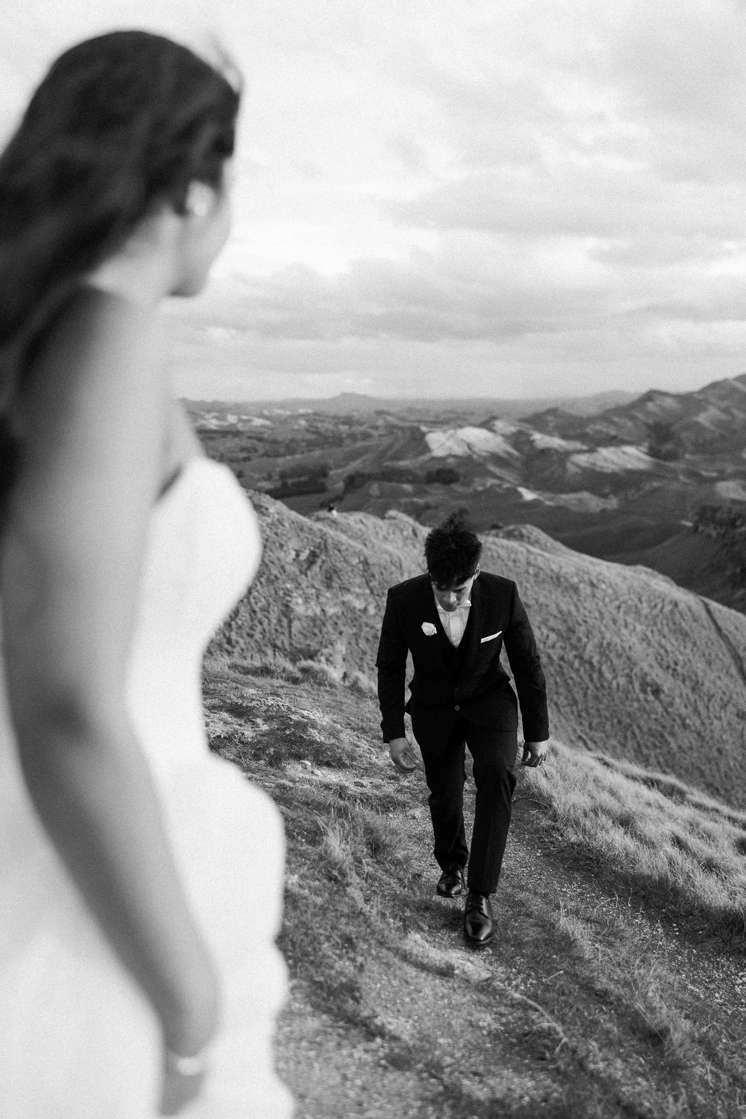 Black and white photo of a groom walking toward his bride on a mountain ridge with scenic valley views.