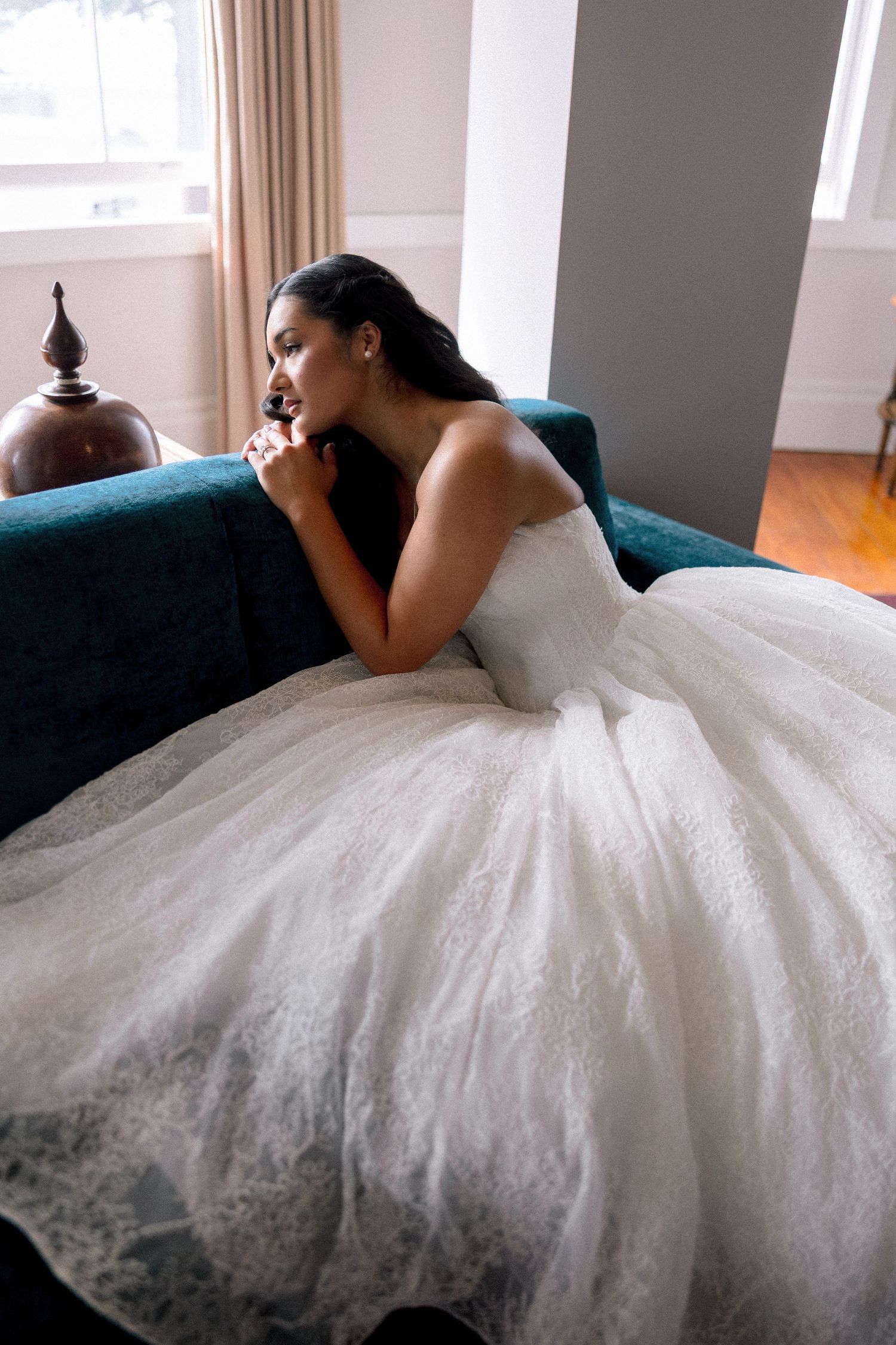 Bride in a stunning white lace ball gown sitting on a teal sofa gazing out a window.