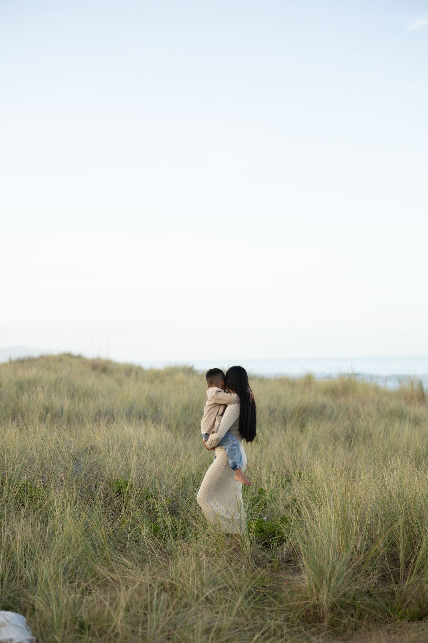 A woman in a beige dress holds a child in a grassy coastal dune landscape under a soft, overcast sky.