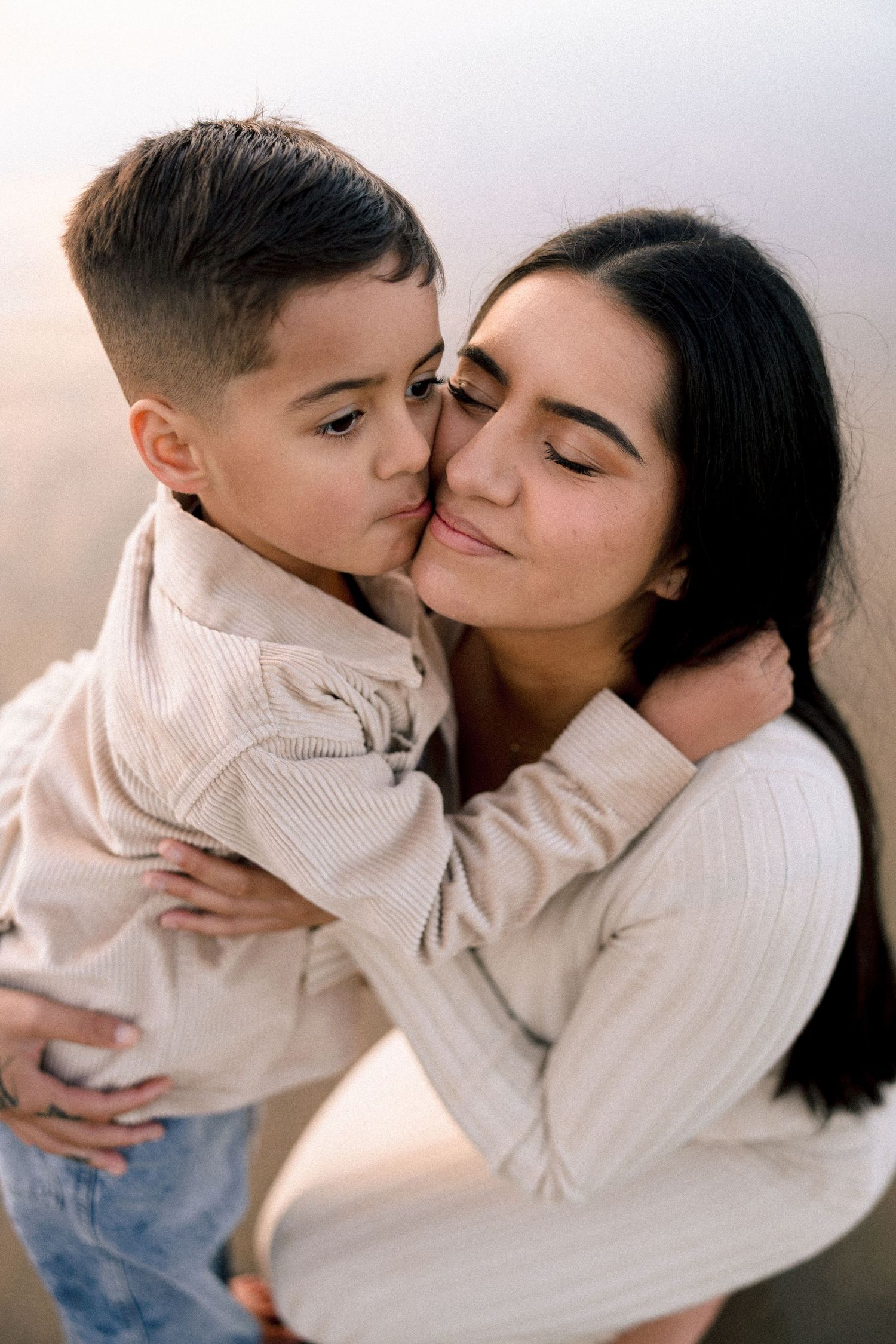 A young boy tenderly kisses his mother on the cheek while they share a warm embrace.