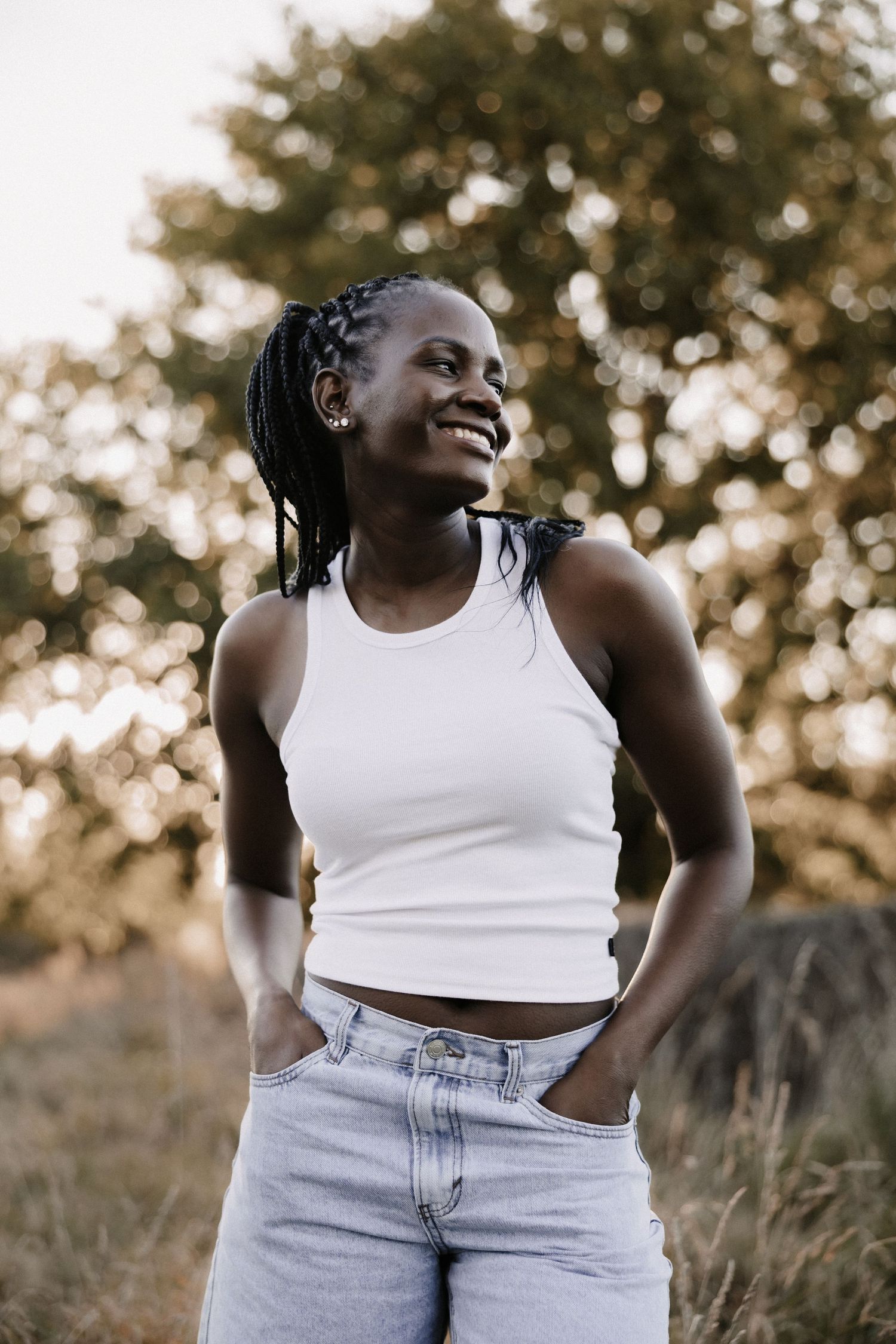 Smiling young Black woman in white crop top and jeans stands outdoors with hands in pockets, trees in background.