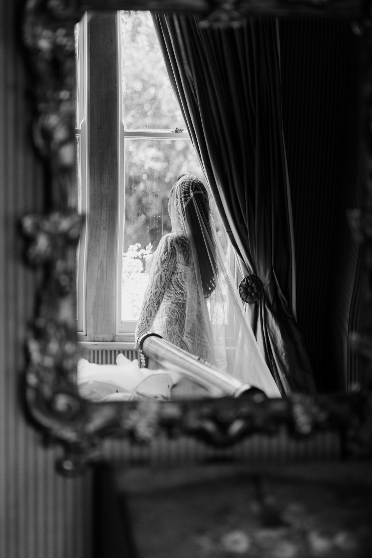 A silhouetted figure stands by a window with curtains in a moody black and white composition.