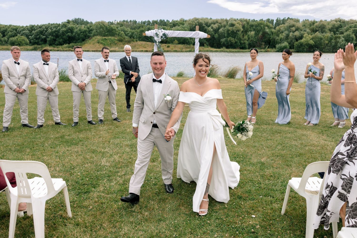 Wedding party in light gray suits and white dresses poses together on a grassy field during an outdoor celebration.