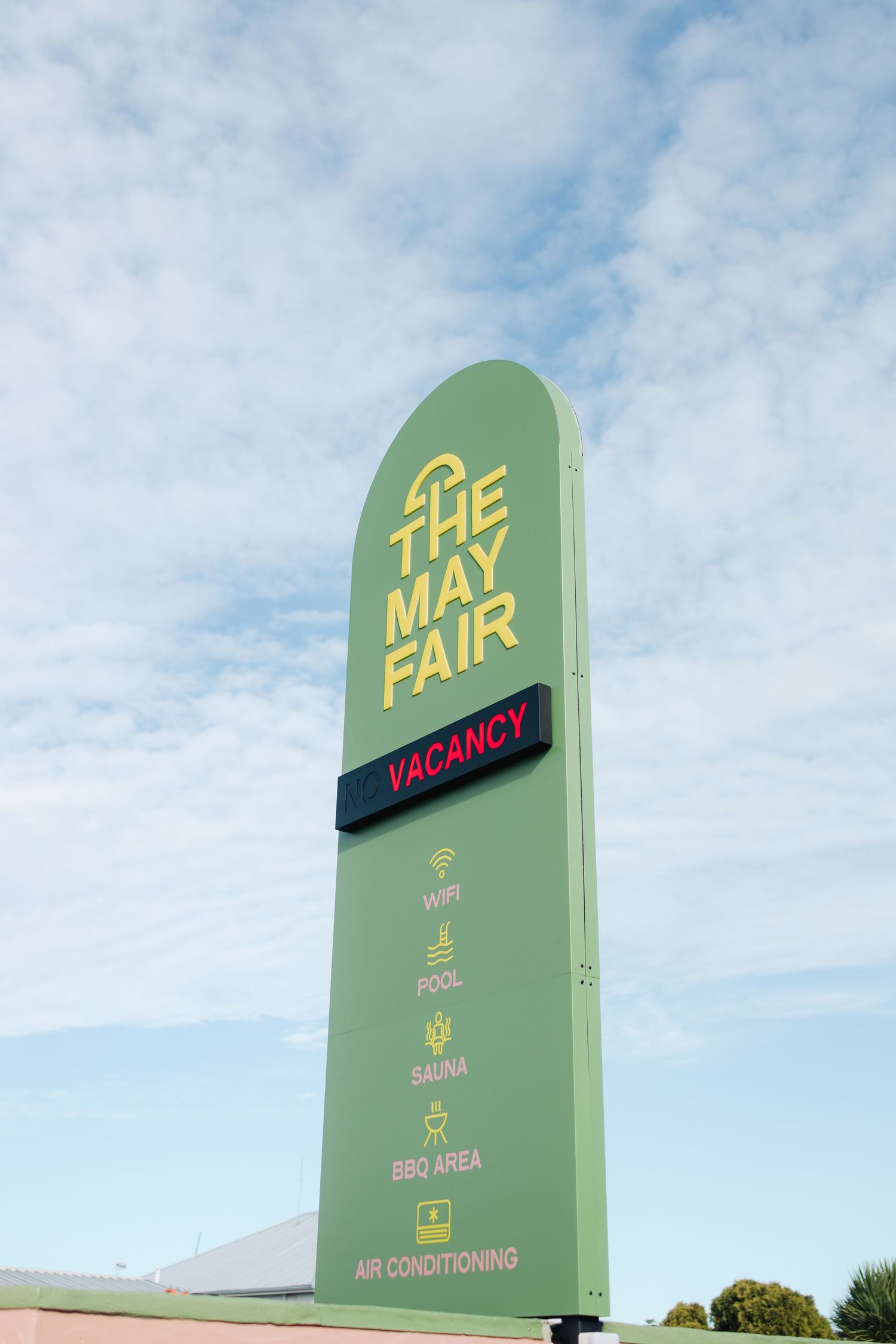 Retro motel sign reading 'The May Fair' with vacancy status against blue cloudy sky.