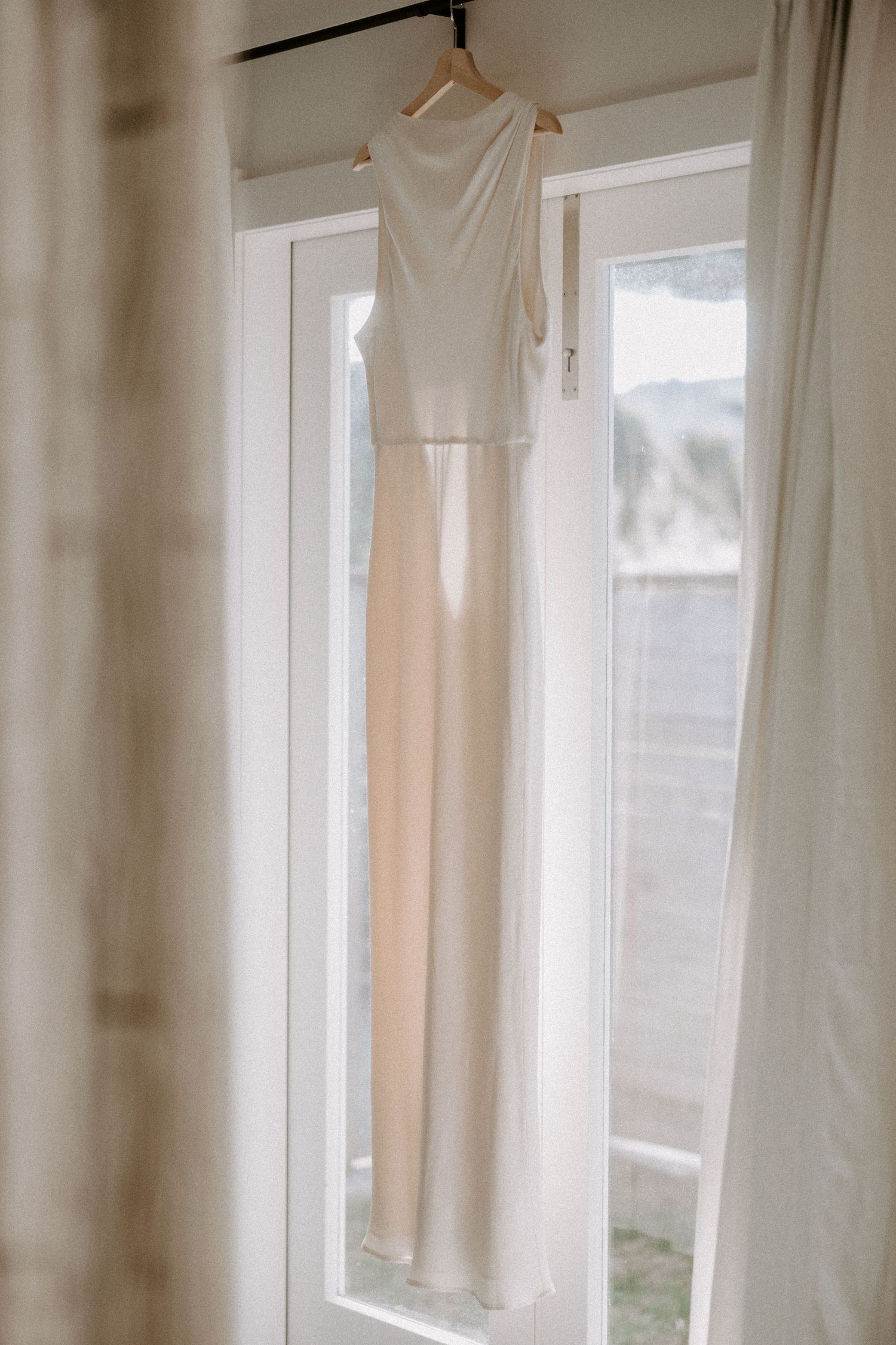 An elegant white wedding dress hangs in front of a bright window, casting soft natural light through sheer curtains.