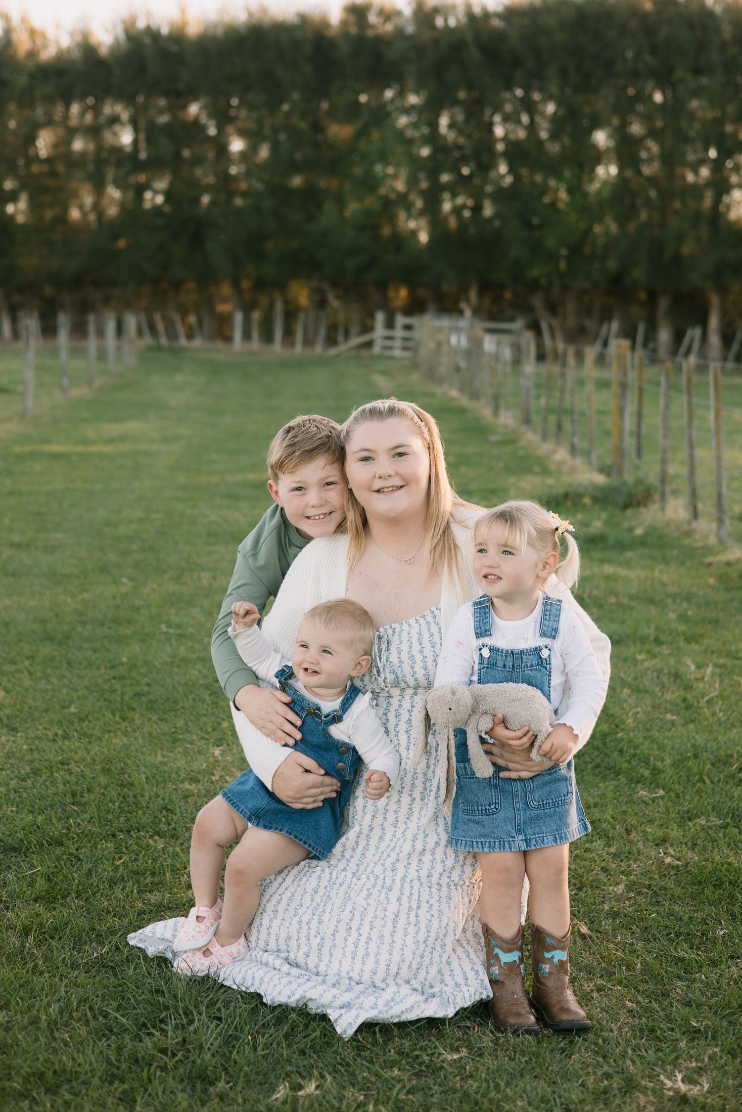 Family portrait in a grassy field with trees, showing group in casual attire sitting together outdoors at sunset.