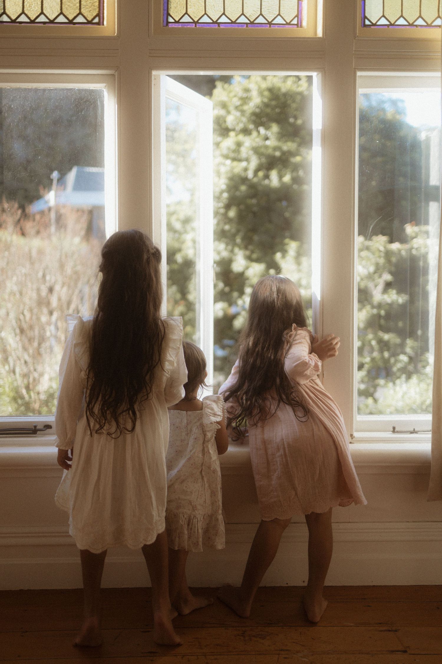 Two children in white dresses stand by a window with decorative stained glass panels, bathed in soft natural light.