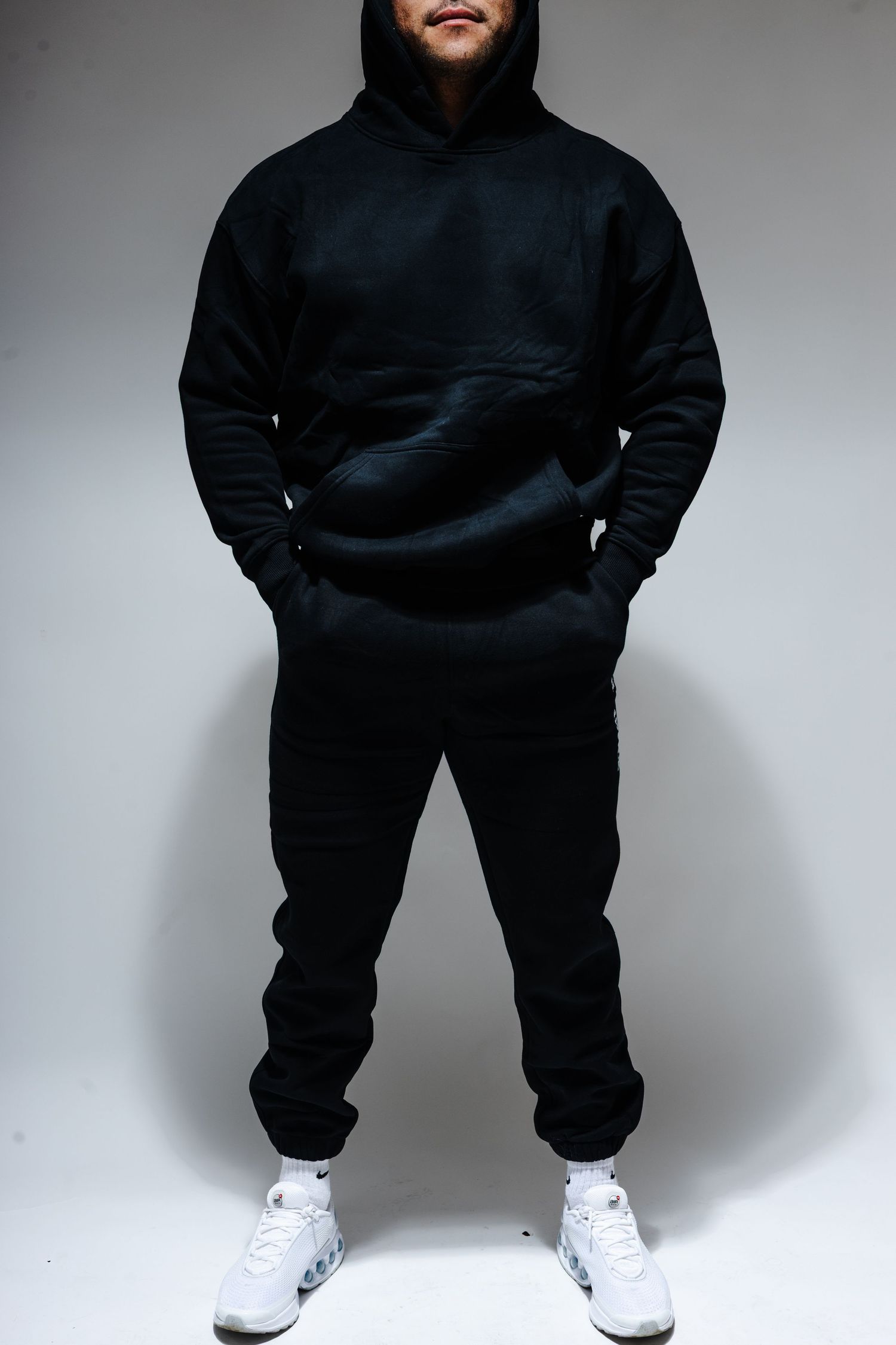 Athletic figure wearing all black hoodie and sweatpants with white sneakers against a gray backdrop.