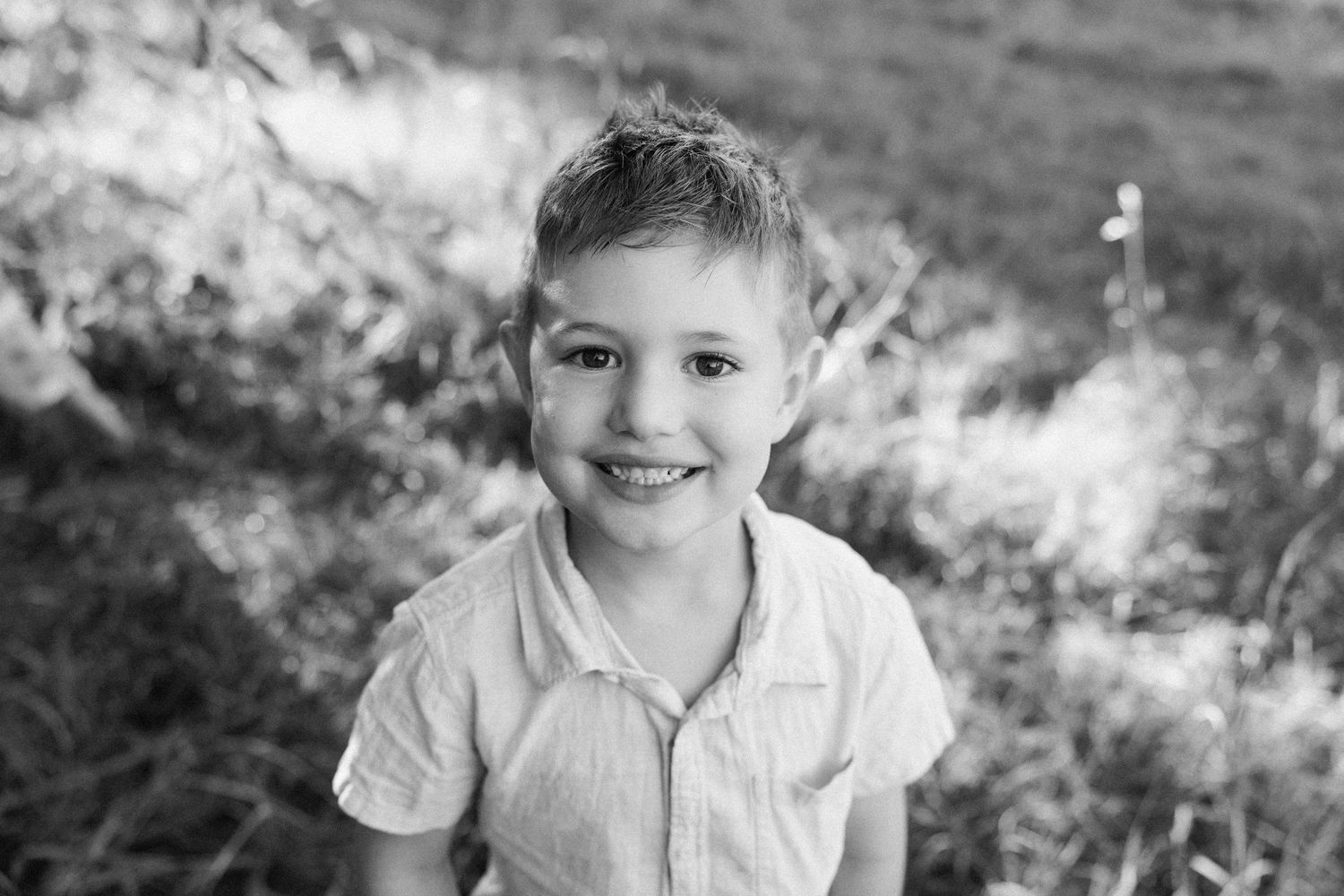 Black and white portrait of a smiling young boy outdoors in a grassy field, wearing a light button-up shirt.