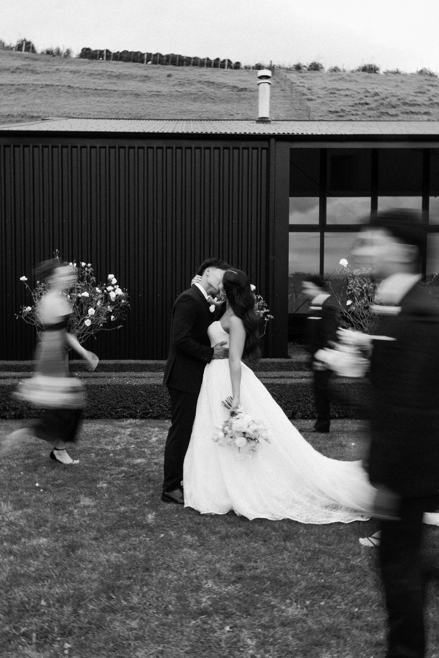 Black and white wedding photo of couple kissing while bridal party walks past in motion blur outside dark barn venue.
