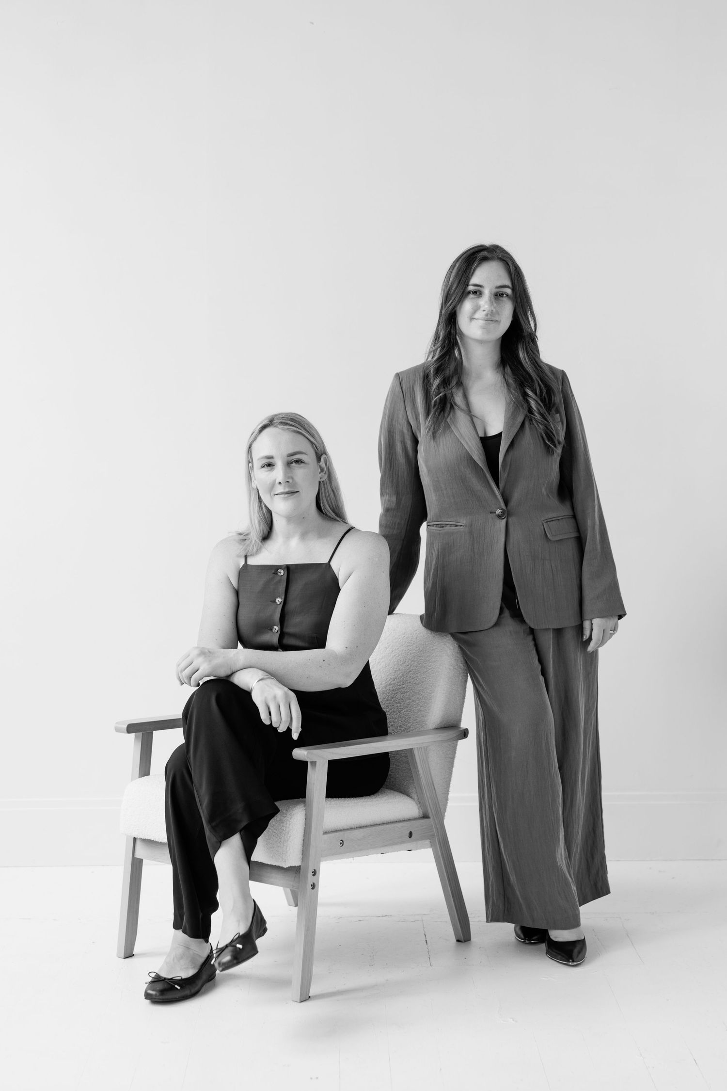 Black and white studio portrait of two women in professional attire posing with one seated in a modern chair.