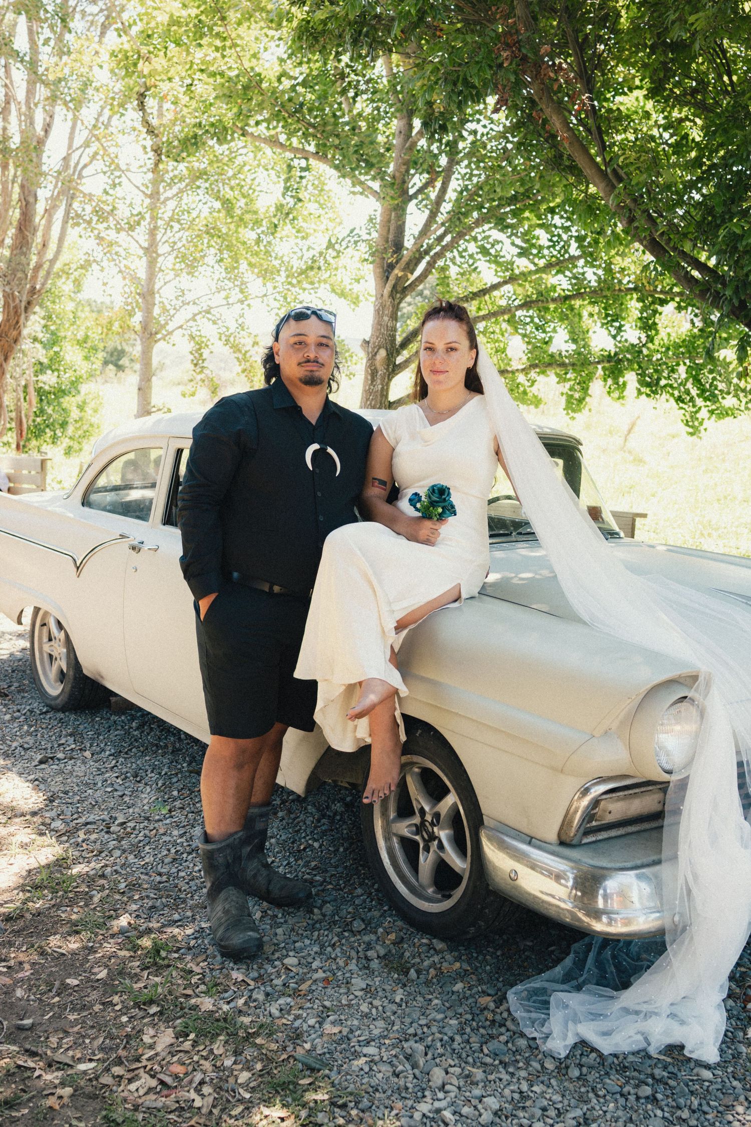 A couple poses with a vintage cream-colored car on a sunny day surrounded by trees.