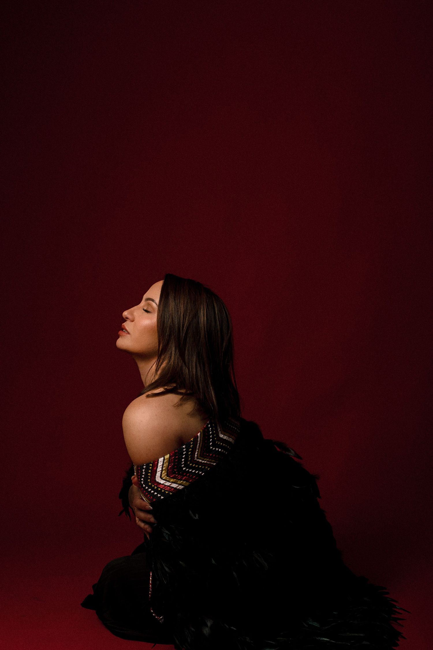 A woman in a black embroidered jacket poses against a deep red background, looking upward with bare shoulders exposed.