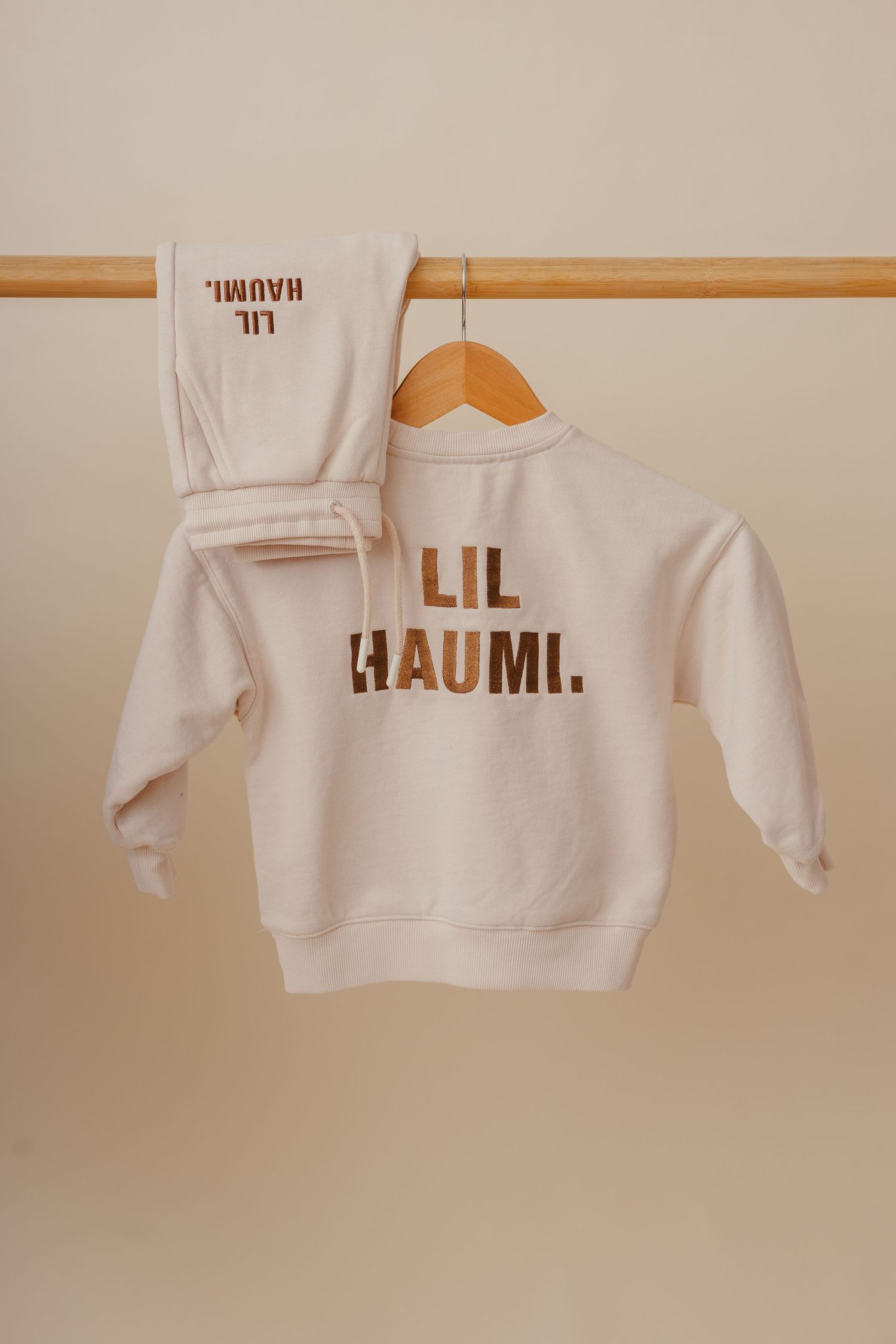 A white sweatshirt with 'LIL RAUMI' text in brown lettering hangs on a wooden clothing rod against a beige wall.