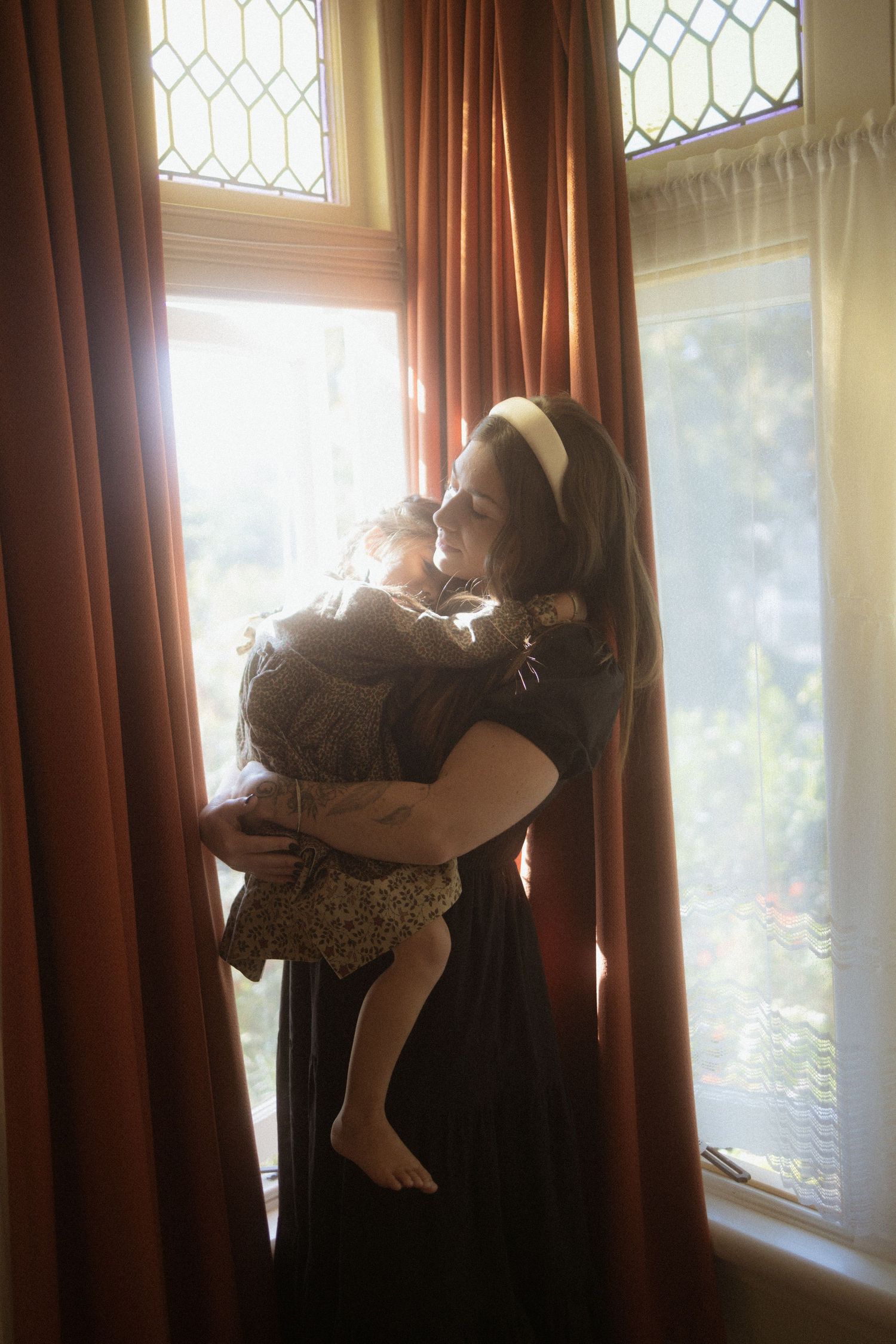 A silhouetted figure holding a child stands near a window with ornate glass and brown curtains creating a dramatic backlight.
