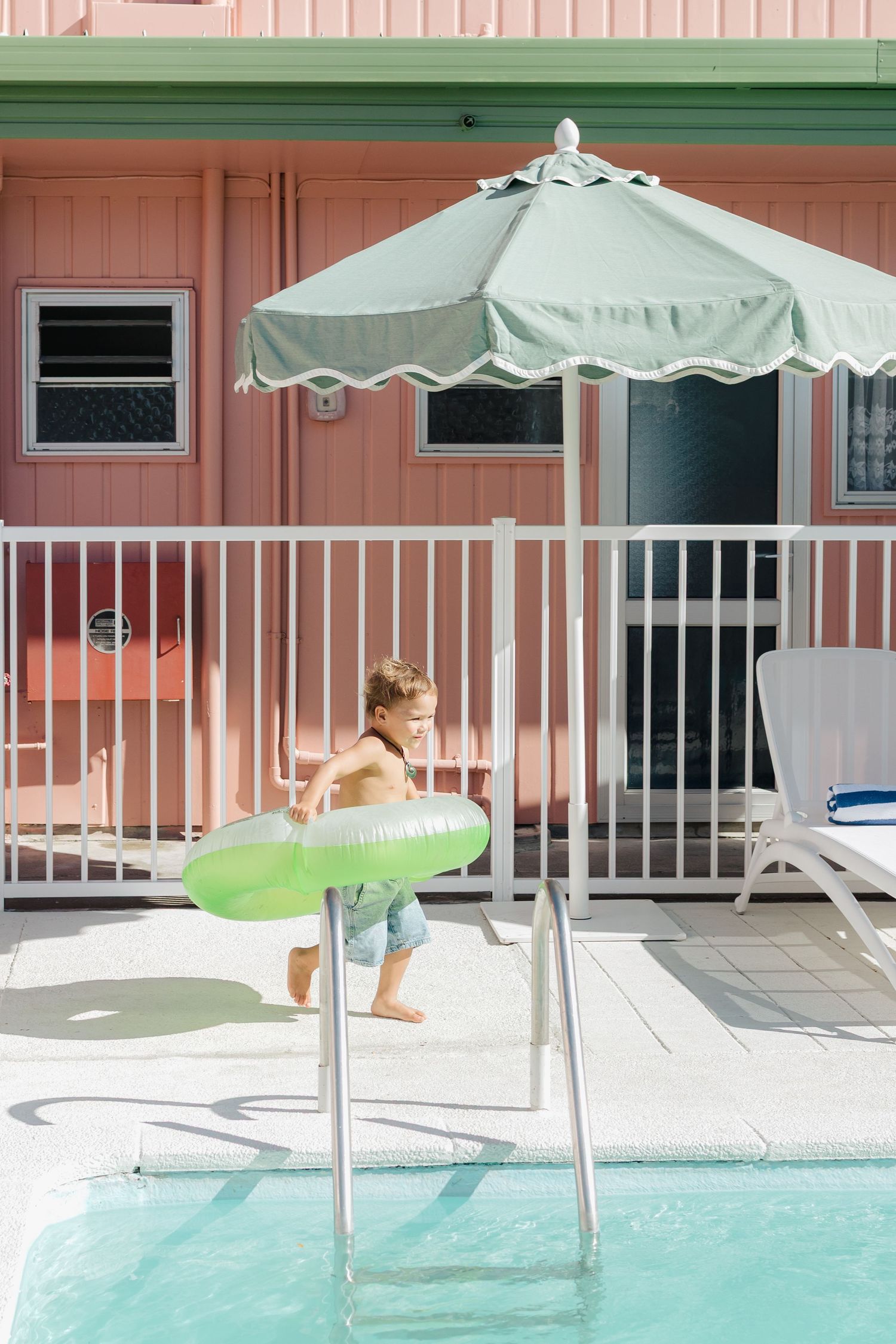Child jumping into swimming pool with green floatie under mint umbrella.