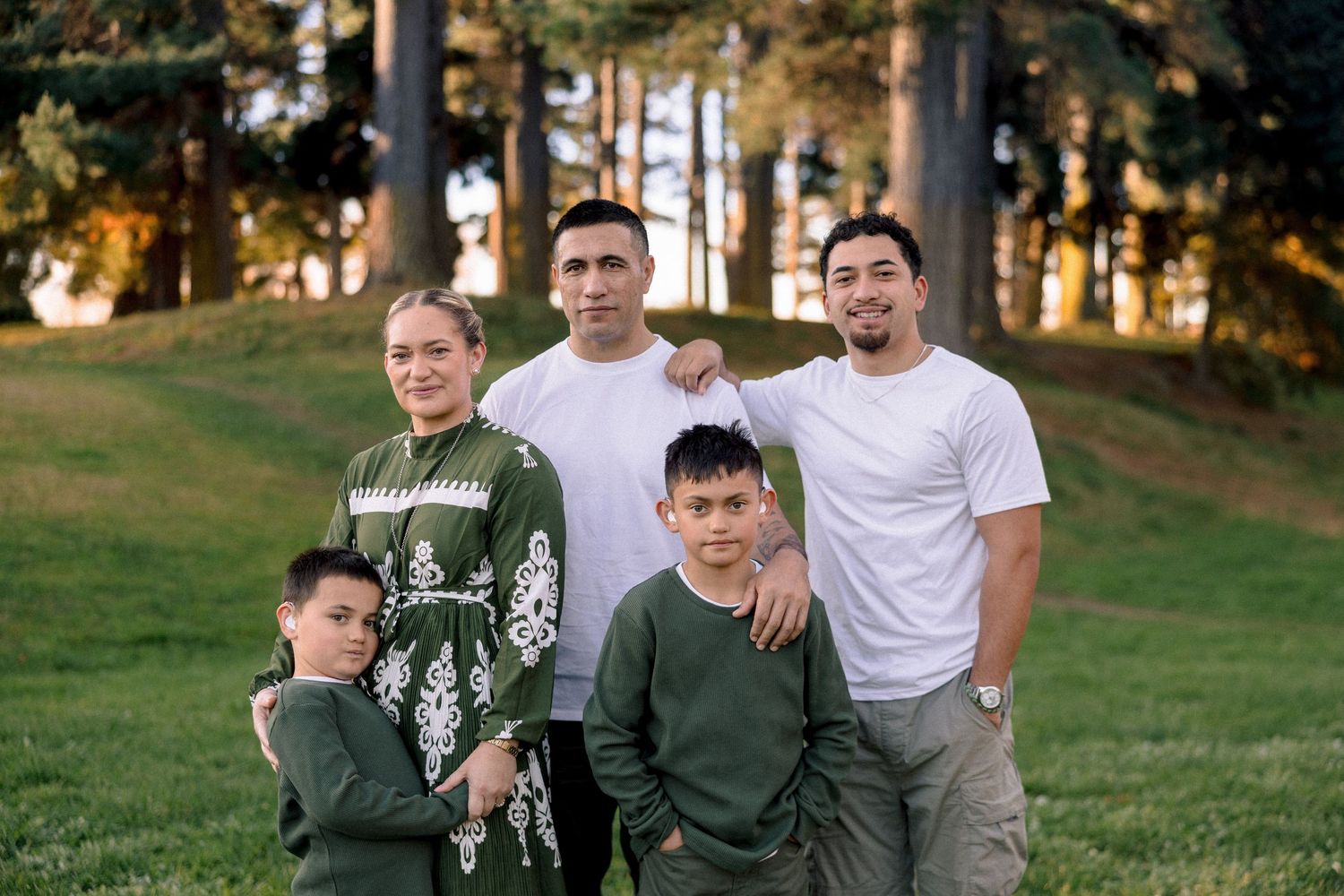 Family of five posing together on a green lawn surrounded by tall trees during golden hour outdoors.
