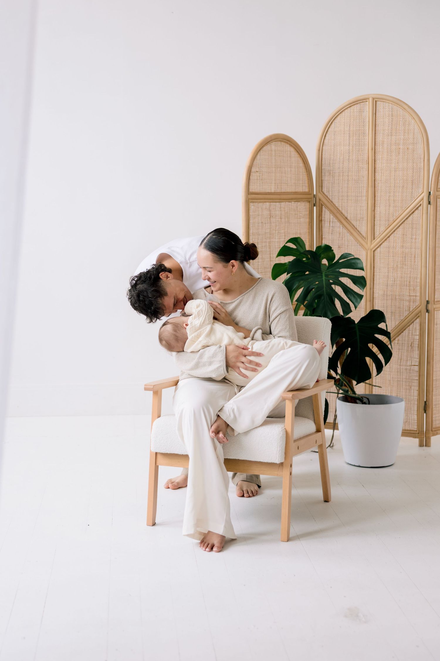 A couple sits in a white armchair, lovingly holding their newborn baby, with a rattan screen and monstera plant behind them.