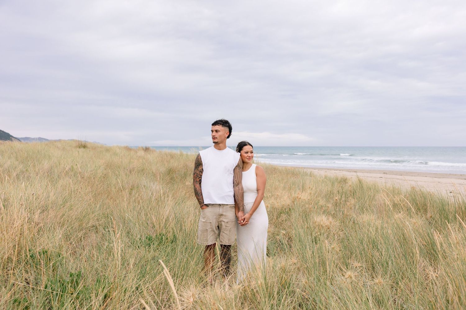 Couple walking hand in hand through tall beach grass near the ocean on a cloudy day.