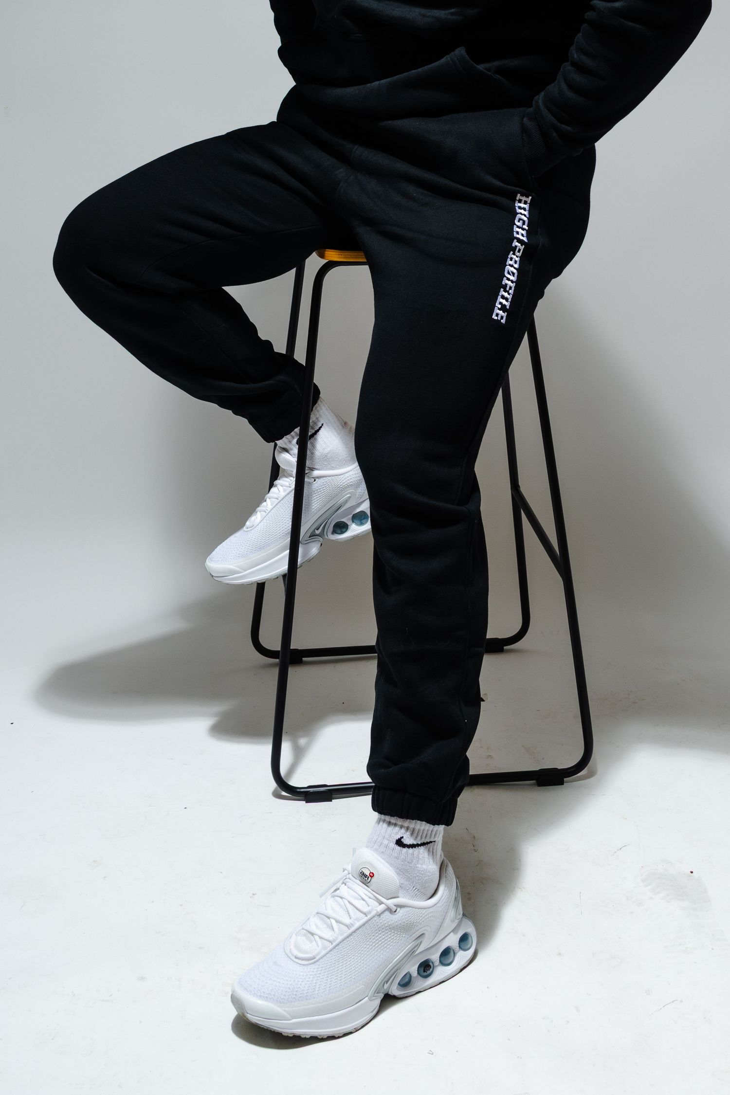 Person wearing black trackpants and white Nike sneakers sits on black metal stool against white background.