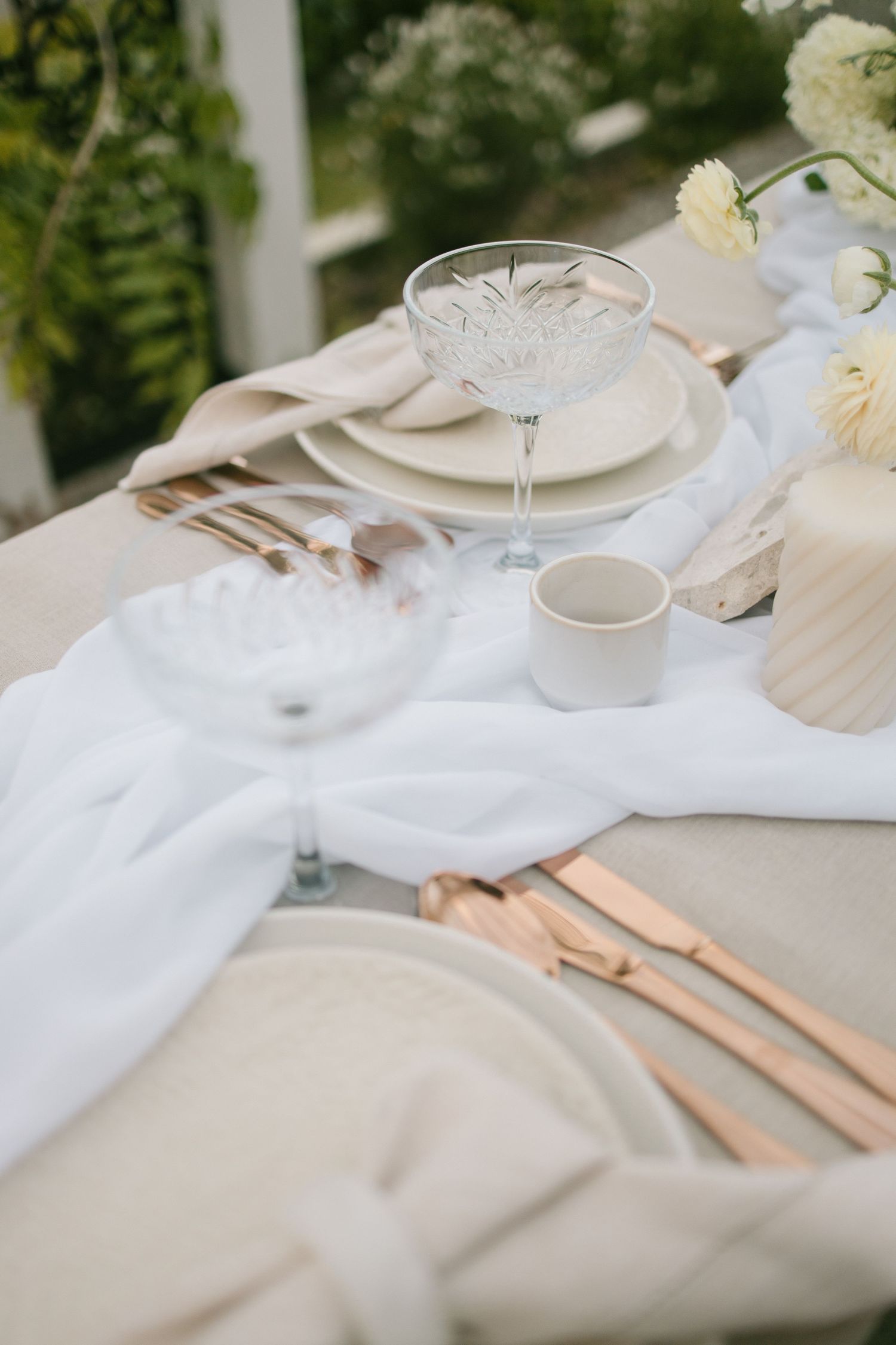 Elegant table setting with vintage crystal glass, white linens, and soft beige plates decorated with white flowers and gold cutlery.