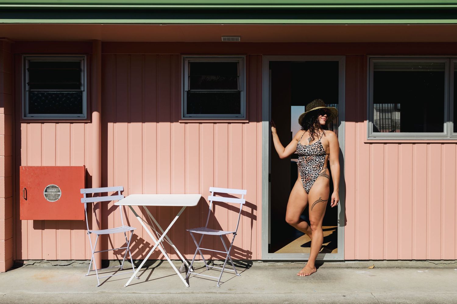 Person in a leopard print swimsuit relaxing at a pink motel with outdoor seating area.