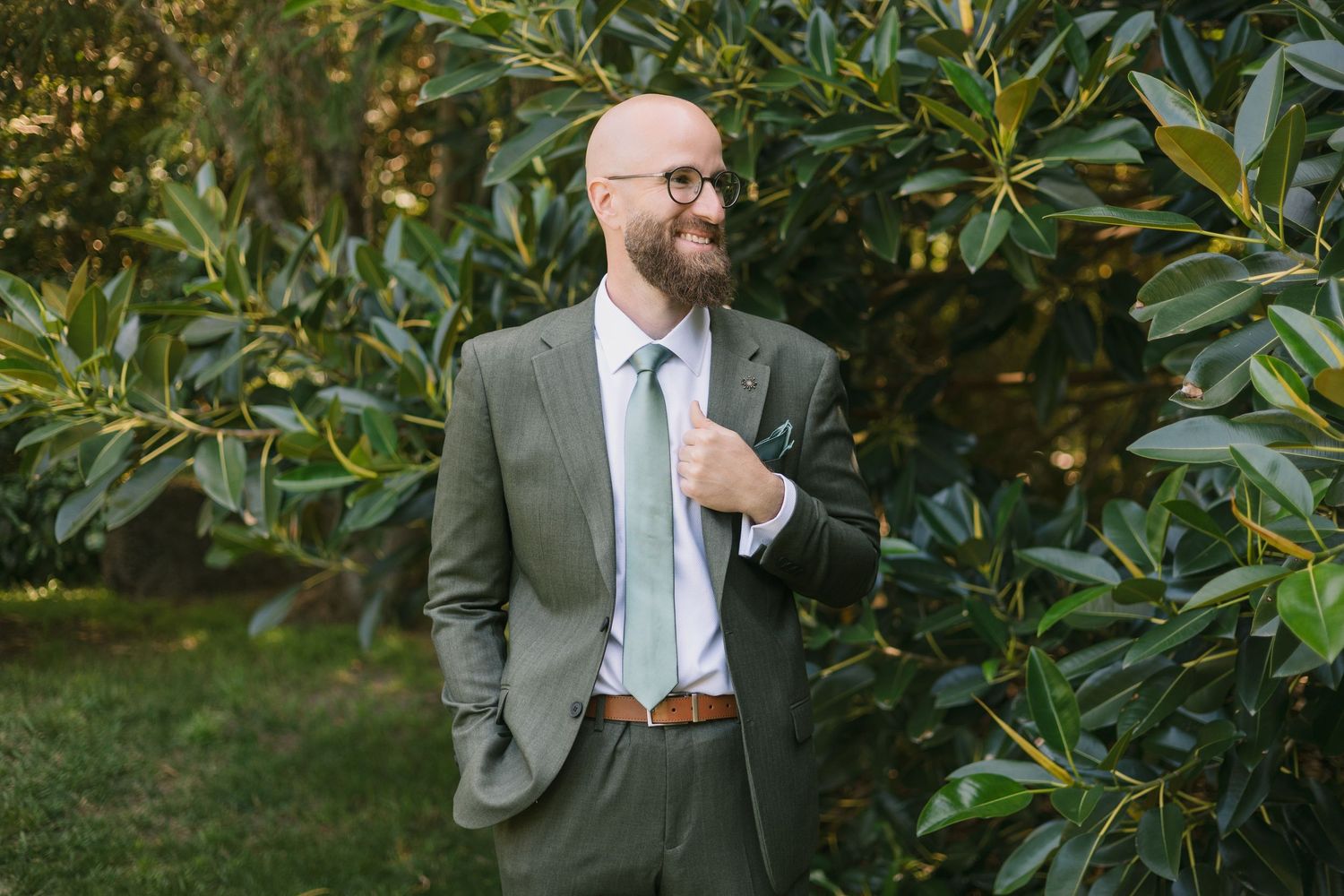 A person in a gray suit and mint tie stands confidently against lush green foliage in an outdoor setting.