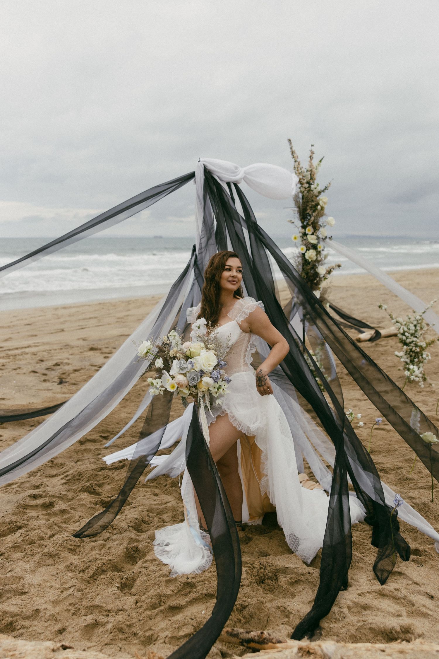 A white wedding dress flows in the wind on a beach with dark fabric and floral decor creating a dramatic arch.