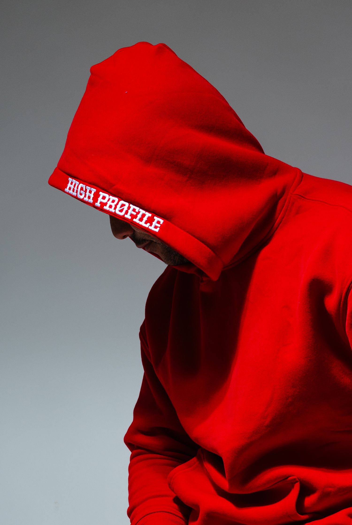 Person wearing bright red hoodie with 'HIGH PROFILE' text against gray background in artistic lighting.