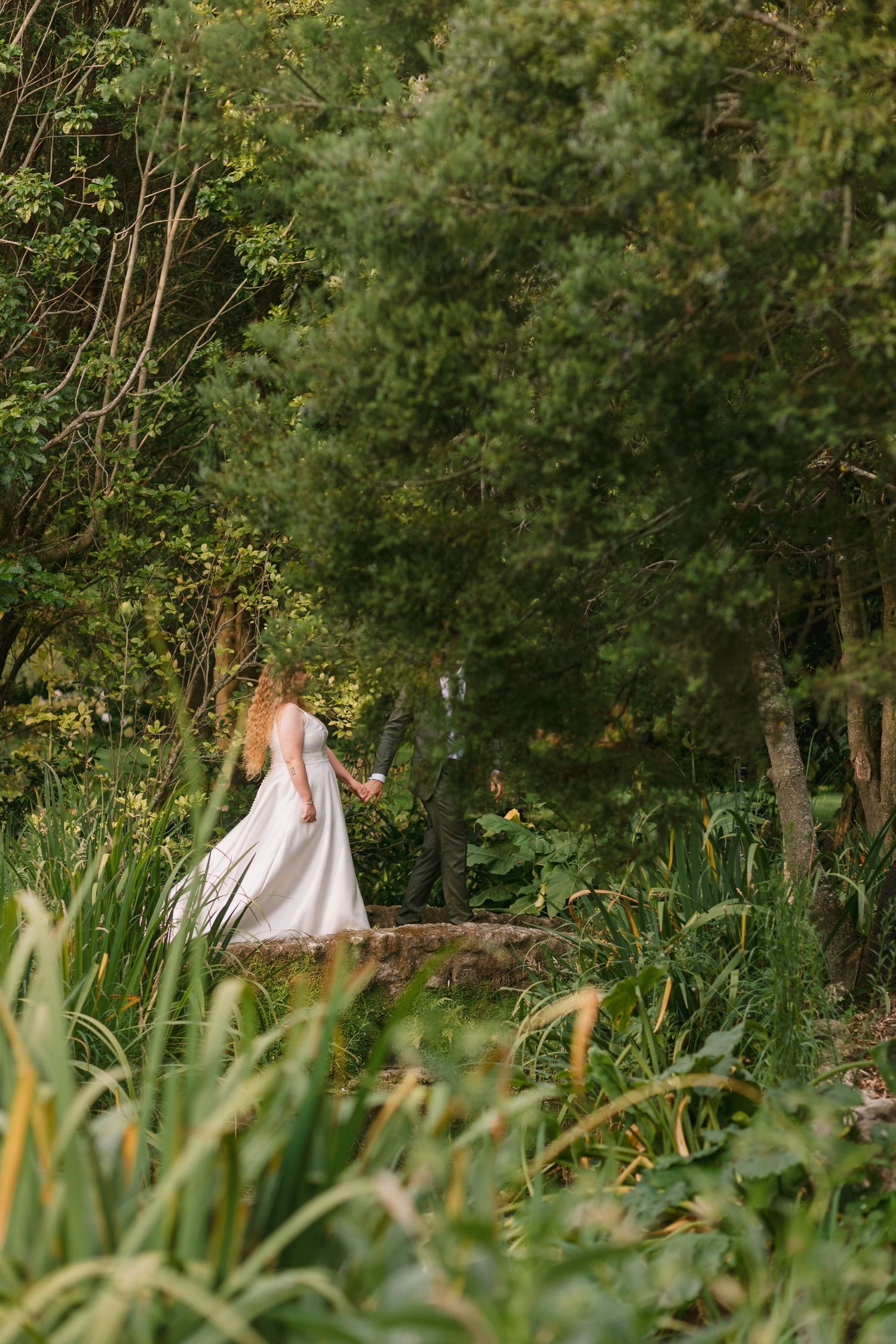 A bride in a flowing white dress walks through a lush garden path surrounded by greenery.
