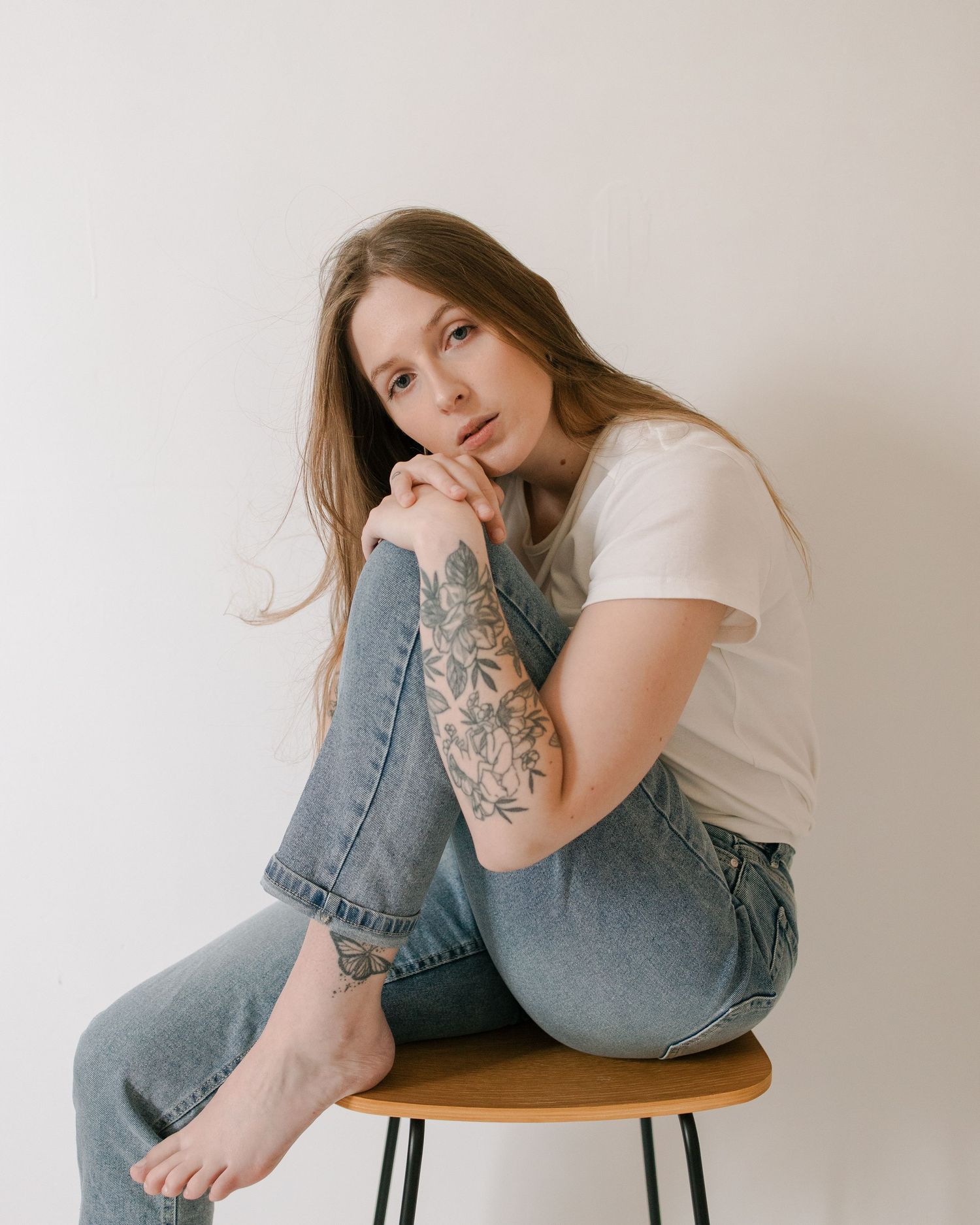 Woman in white t-shirt and jeans sits on wooden stool against white wall in artistic portrait series.