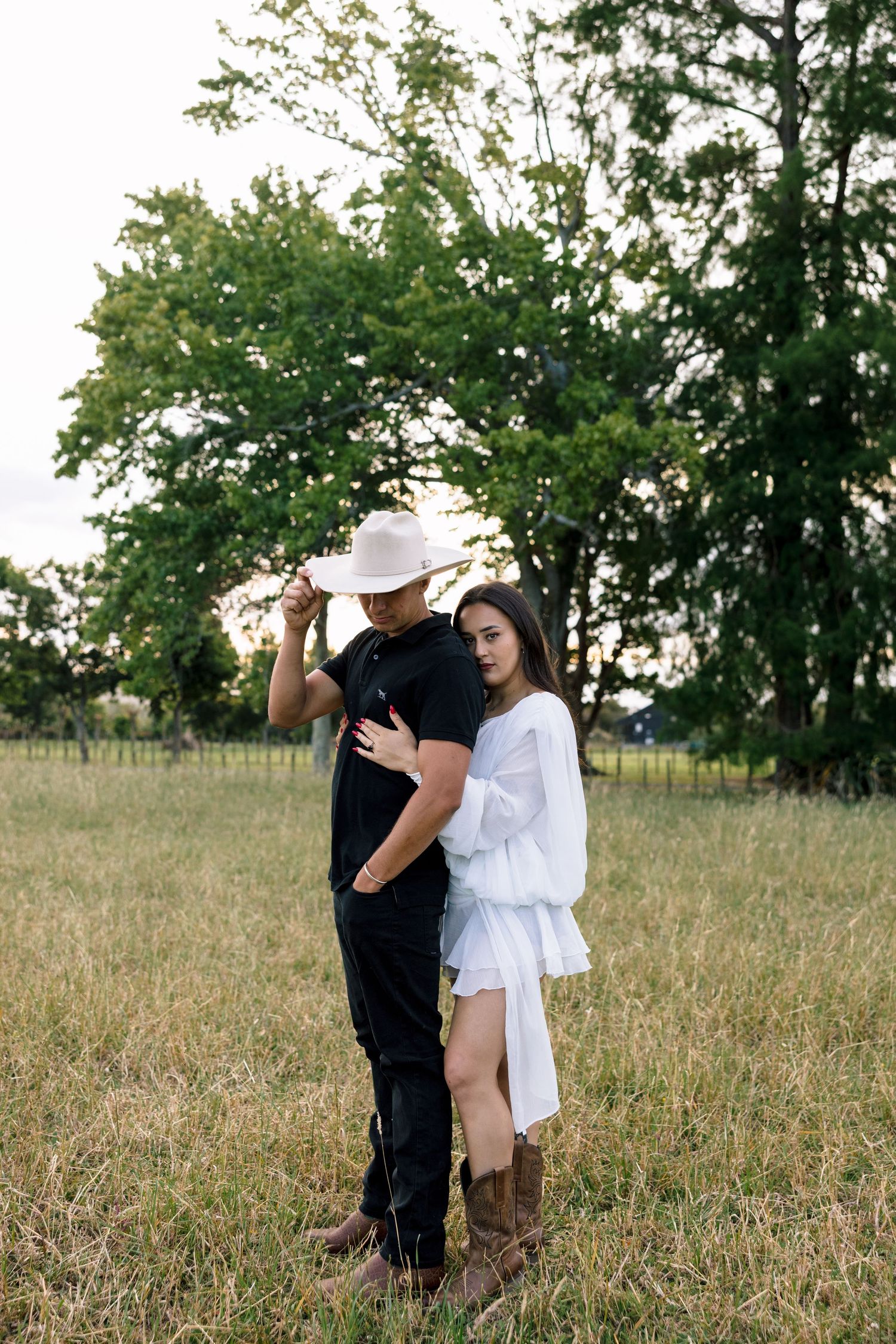 A romantic country scene with a couple in contrasting outfits standing together under a large tree.