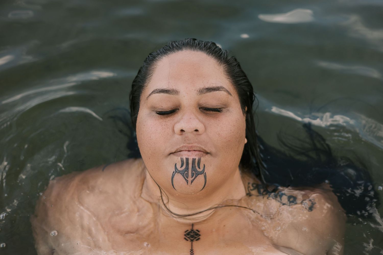 Person floating in dark water with closed eyes and a chin tattoo.
