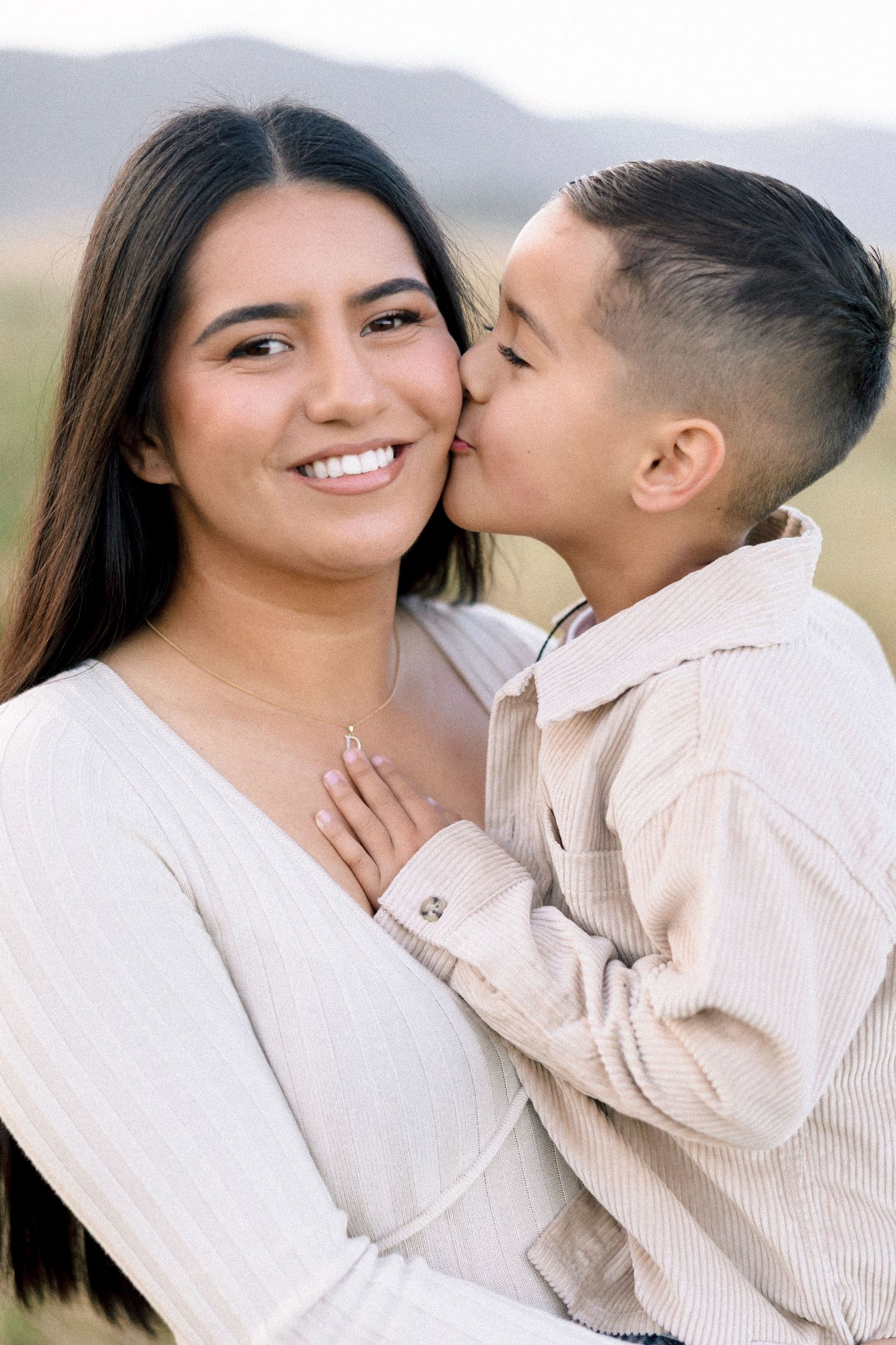 A young boy kisses his smiling mother on the cheek outdoors in a field.