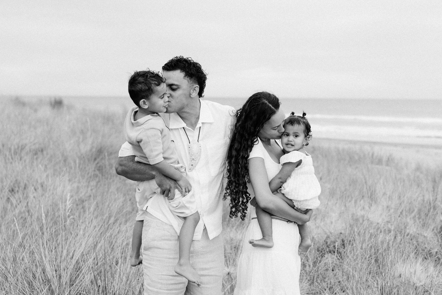 A couple in white outfits embraces lovingly in a grassy field by the beach during a romantic photoshoot.